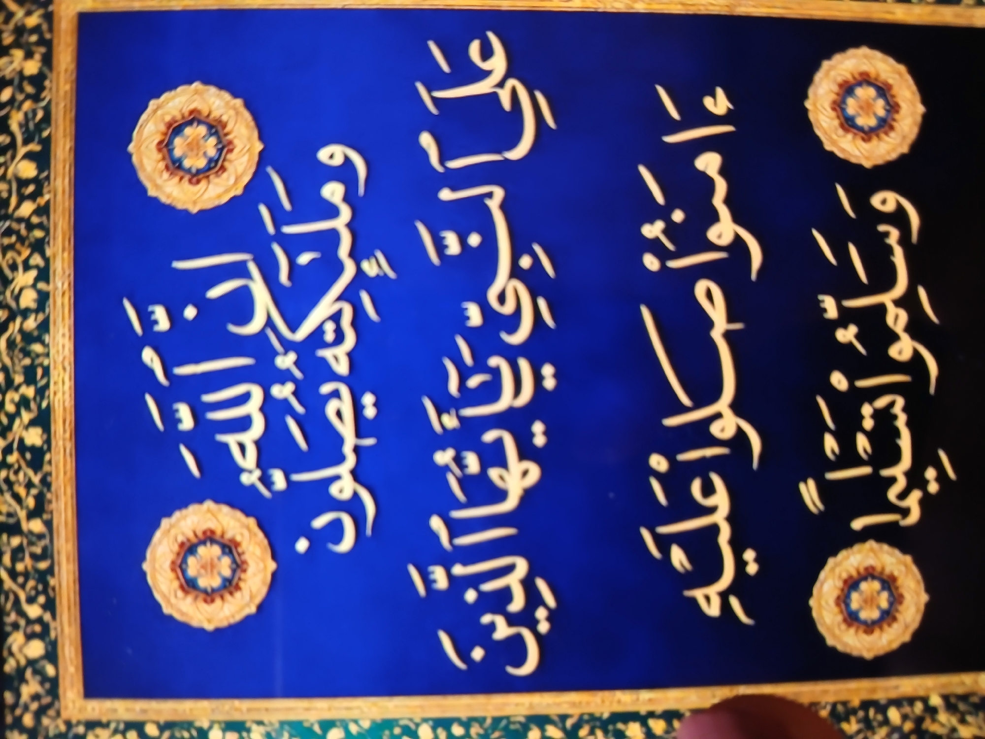 The image shows a framed piece of art, potentially calligraphy, on a dark blue background with a decorative floral border. The calligraphy, rendered in a light color, is arranged in vertical columns with ornamental rosette designs placed in the four corners of the frame. The artwork is framed with a thin golden trim. The overall scene has a rich, ornamental aesthetic. The exact location in Bengaluru, India, is not discernible from the image.