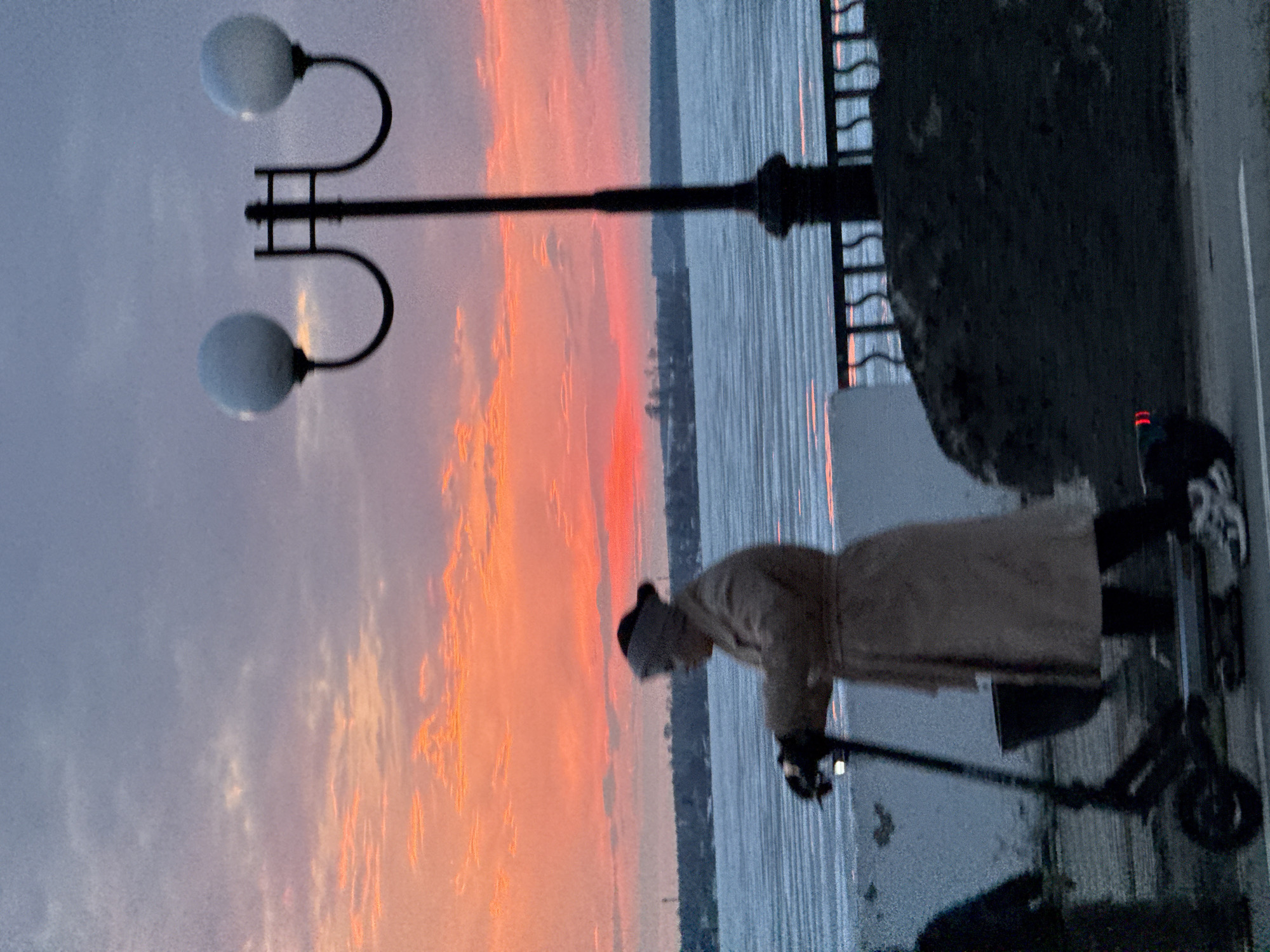 A person is riding a scooter on a paved surface along the edge of a body of water, likely a reservoir or wide river, with a dramatic sunset in the background. The location is possibly in Votkinsk, Russia.
The person is wearing what appears to be a light-colored coat and a dark head covering. They are standing on the scooter with their hands on the handlebars. The scooter is small and dark-colored.
To the left of the person is a street light with two globe-shaped lights. Behind the scooter is a low retaining wall with metal railings. The body of water has a vertically striated appearance, suggesting either waves or a photographic artifact.
The sky is filled with a sunset displaying hues of orange, pink, and blue.