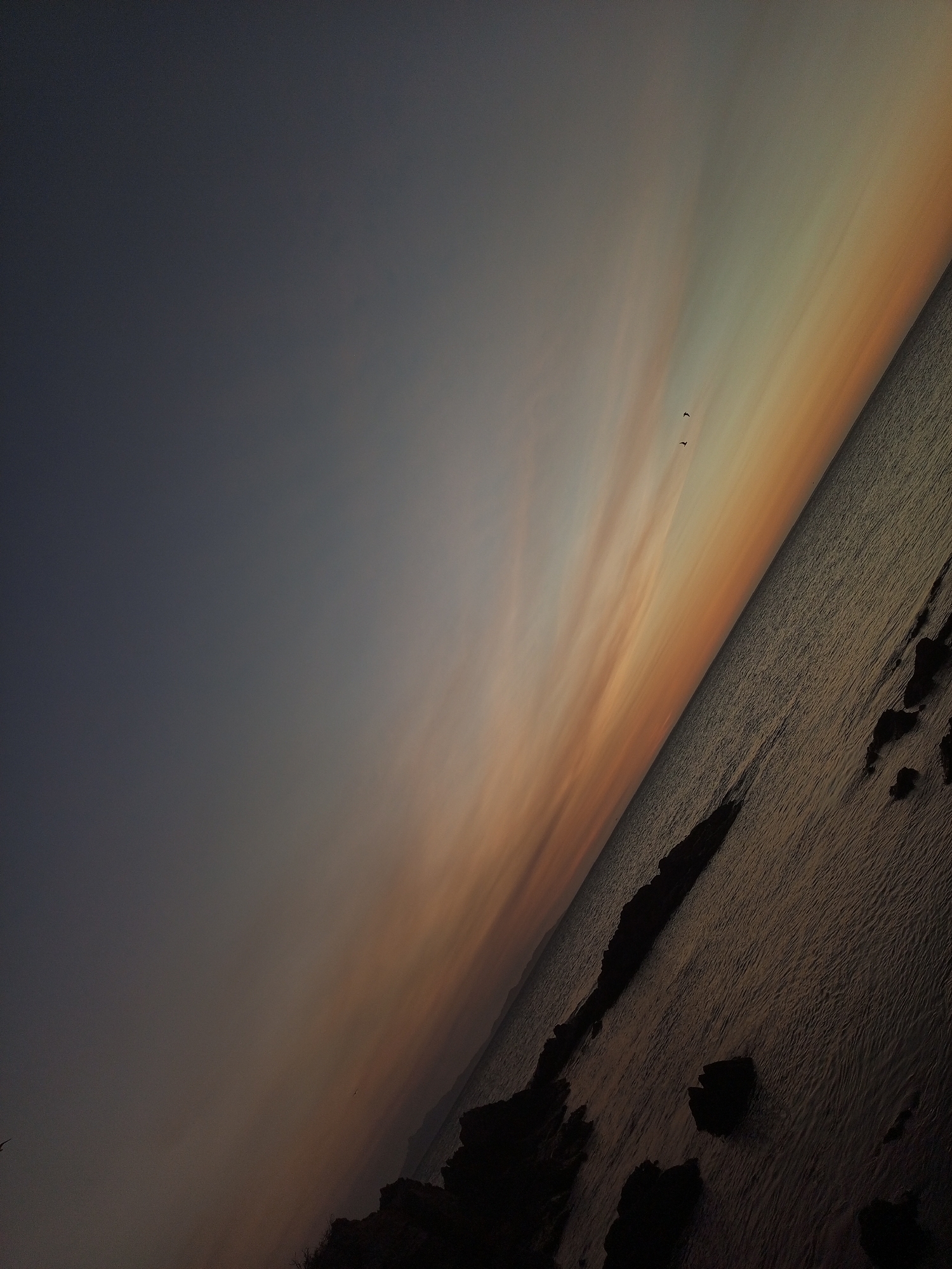 The image depicts a body of water, possibly the ocean, divided by an angled horizon. The sky transitions from a dark blue-gray to a gradient of yellow, orange, and peach hues near the water's edge. Two birds are seen silhouetted against the lighter portion of the sky. Jagged dark rocks are visible along the water's edge. The photo may have been taken in Juan Griego, Venezuela.