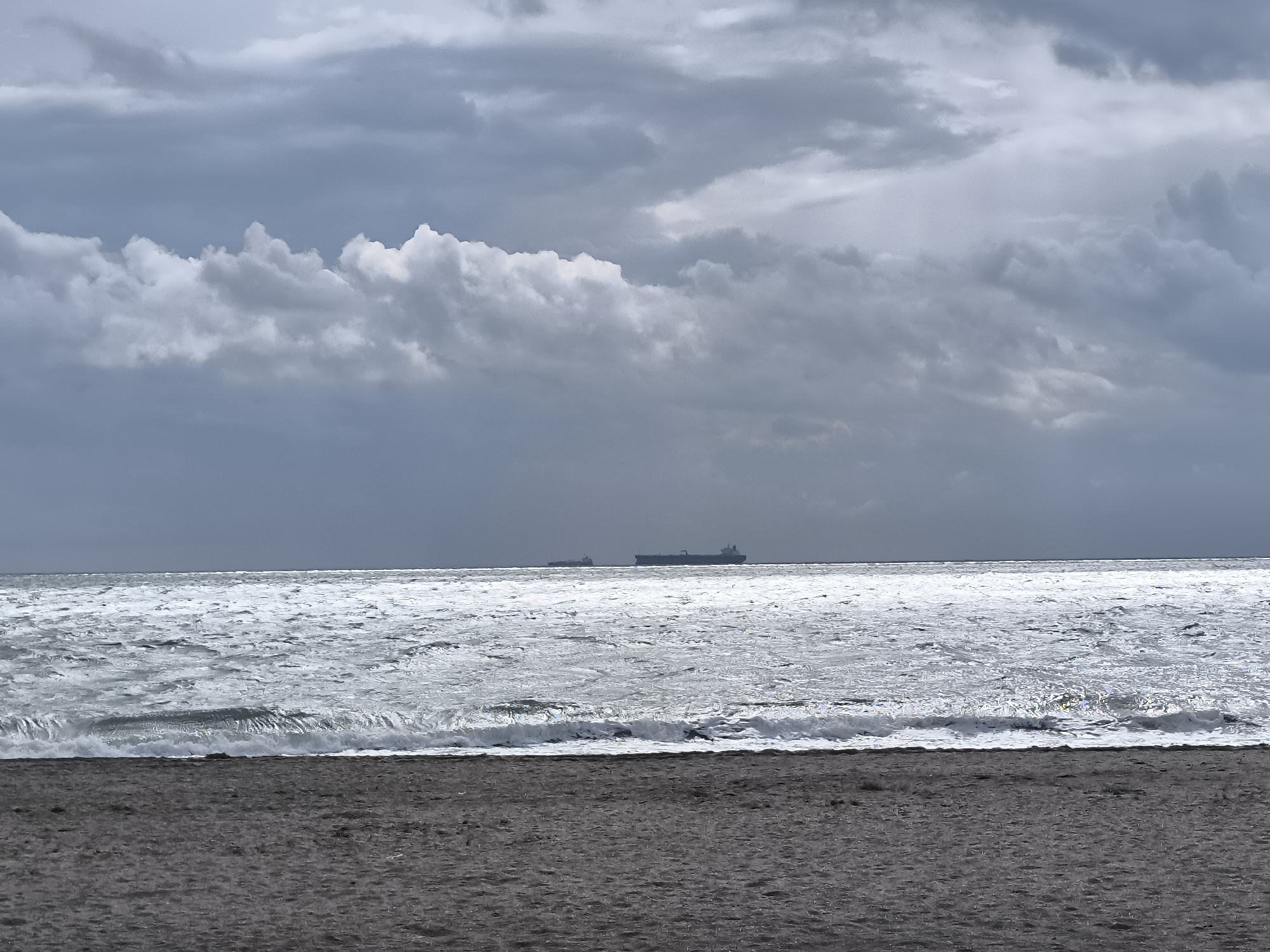 The image depicts a beach scene, likely in Burgas, Bulgaria. The foreground consists of dark sand. Beyond the sand is a turbulent sea with white-capped waves. In the distance, two ships are visible on the horizon. The sky is overcast with heavy, gray clouds. The overall impression is of a cloudy, windy day at the beach.