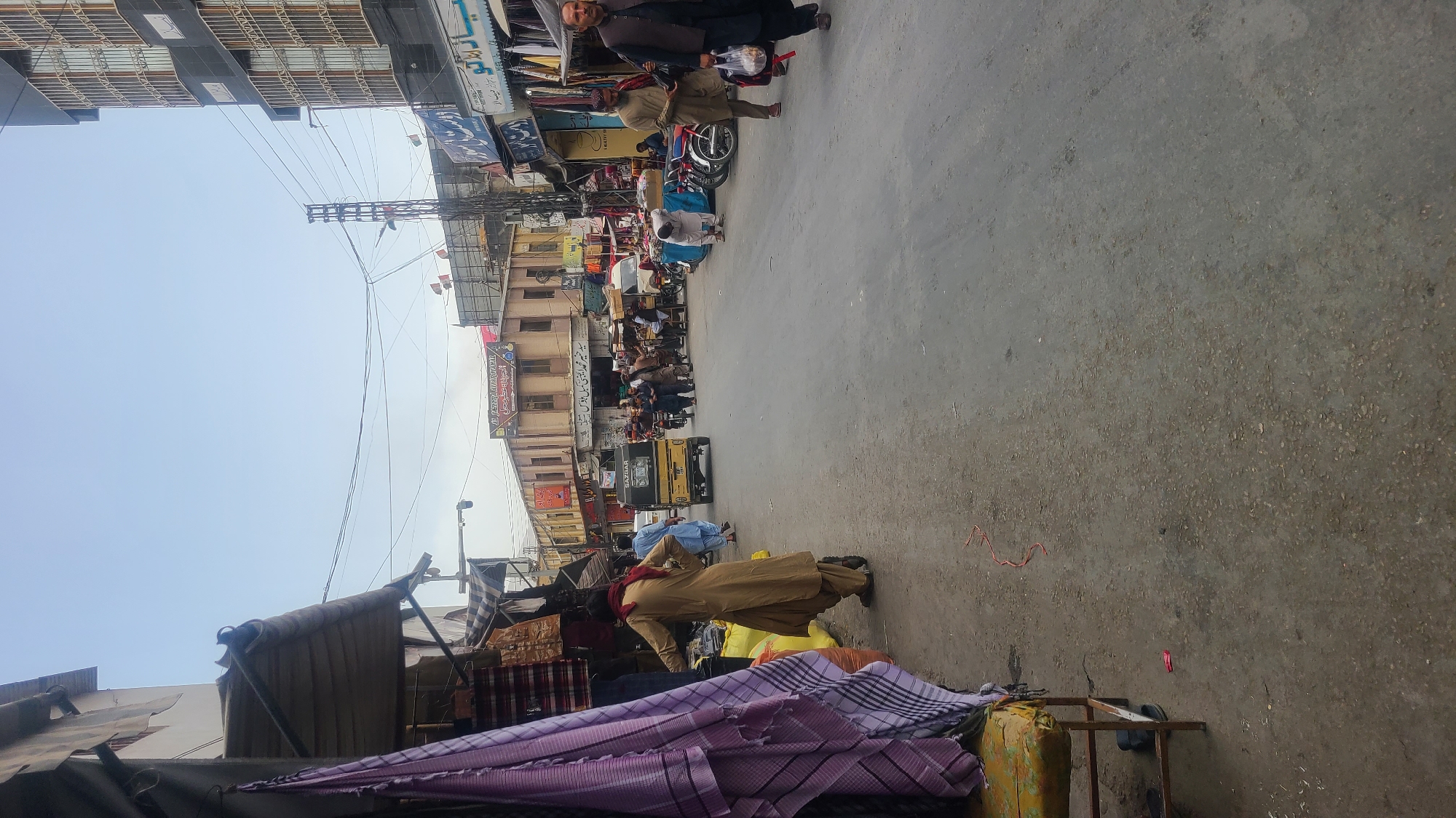 The image depicts a street scene in Quetta City Tehsil, Pakistan. The view looks down a busy commercial street with buildings on either side. The street has a mix of pedestrians, motorcycles, and a yellow auto-rickshaw. Several men are visible, dressed in traditional Pakistani clothing. A person wearing a tan shalwar kameez is standing near some textiles displayed on a table. Shops with awnings and signage are visible along the sides of the street. Power lines crisscross overhead. The sky is bright and mostly clear.