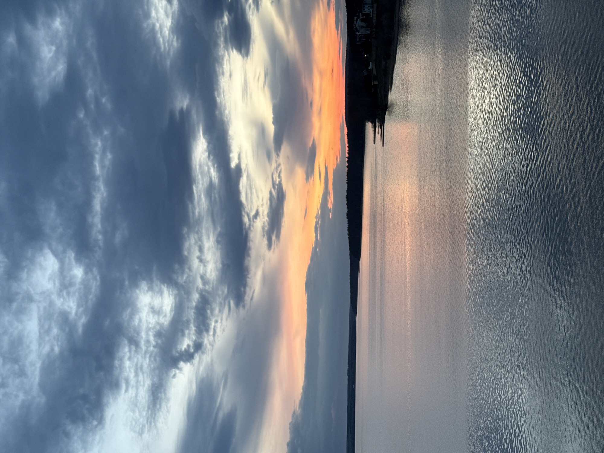 The image depicts a body of water, likely a lake or wide river, adjacent to a shoreline with trees and some buildings. The sky is filled with clouds, ranging from dark and stormy on the left to lighter with orange and yellow hues near the center, suggesting sunset or sunrise. The water surface reflects the sky's colors. It is not possible to confirm the location in Votkinsk, Russia without additional information.