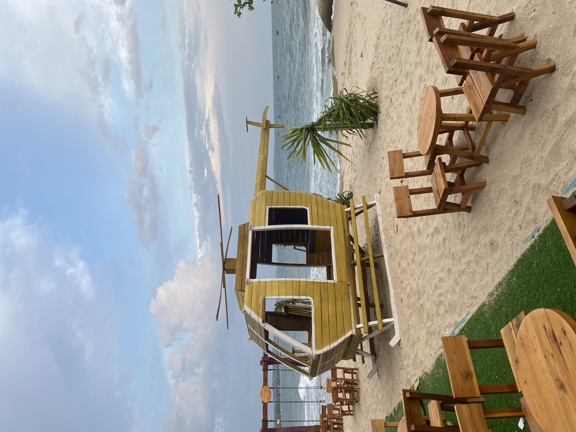 The image shows a beachside setting in Phu Thuy Ward, Vietnam. A yellow helicopter-shaped structure sits on the sand. Tables and chairs are arranged on the beach. The ocean is in the background. The sky is cloudy. The scene appears to be a restaurant or leisure area.