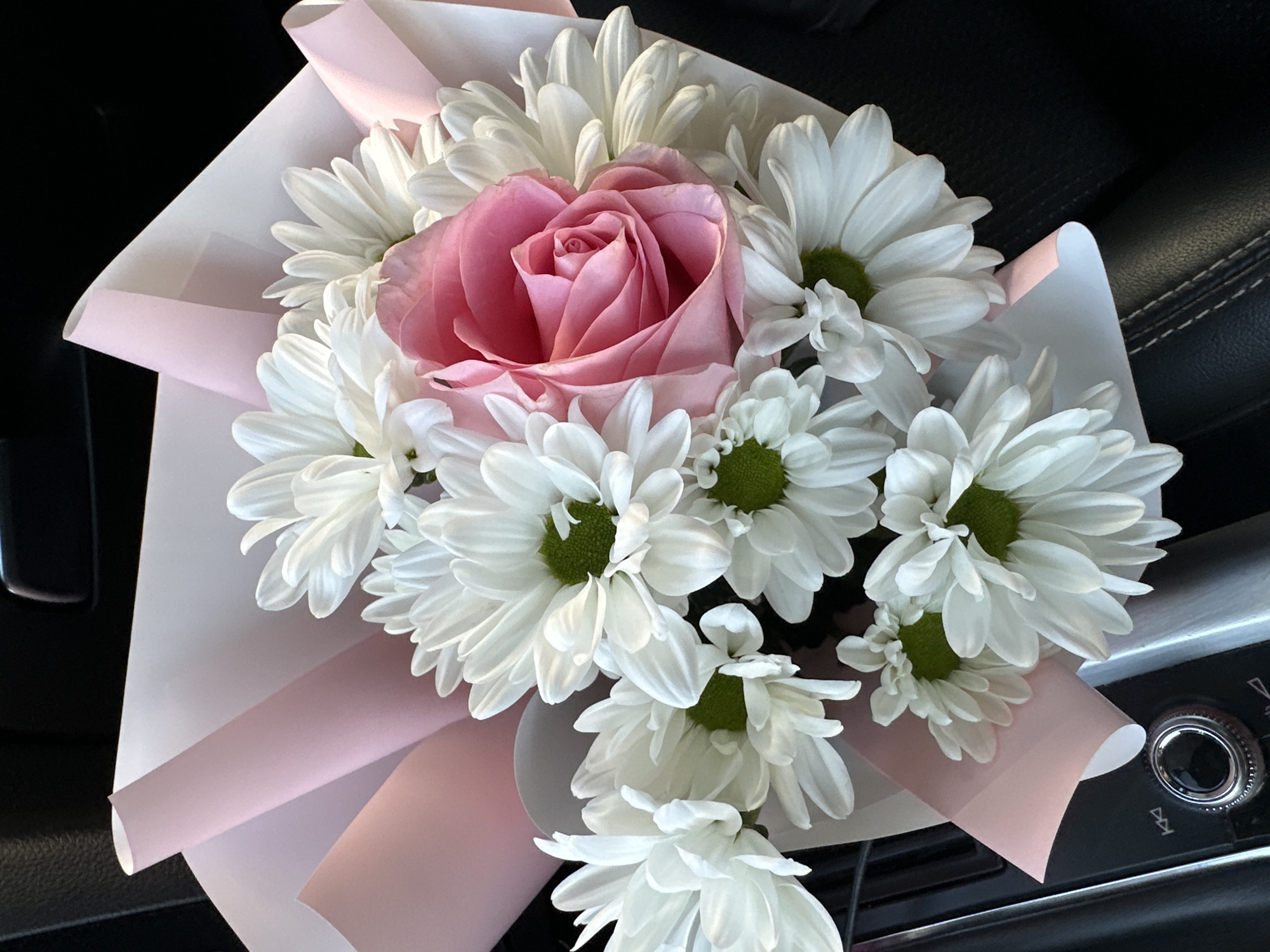 A close-up shows a bouquet of flowers, featuring a large pink rose surrounded by white daisies with green centers, wrapped in light pink paper. The bouquet is positioned on a dark surface, possibly a car interior, with a portion of the dashboard and stitching visible. A car entertainment system with a large dial is visible in the bottom right corner. The location appears to be inside a car, likely in Sofia, Bulgaria, though there is no concrete evidence to support this. No people are present.