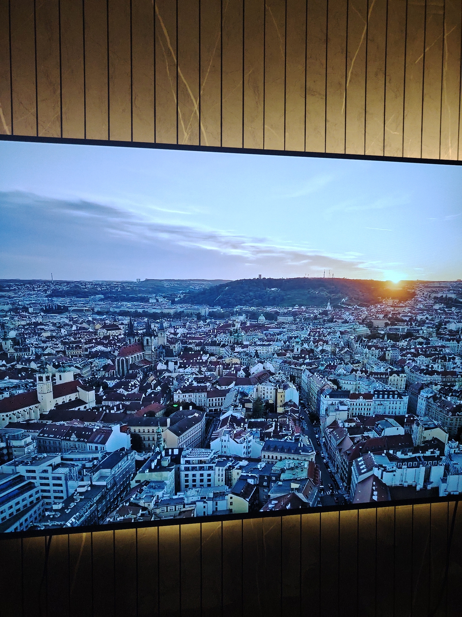 A large rectangular screen displays an aerial view of a densely populated European cityscape at sunset/dusk. The sky above the city is a light blue with scattered clouds, transitioning to an intense orange-yellow glow on the far right horizon, indicating the setting sun. The city below features numerous historic buildings, characterized by pitched reddish-brown roofs, a mix of architectural styles, and several prominent churches with spires and towers. A significant tree-covered hill is visible in the background on the right, with distant structures on its summit, consistent with Petřín Hill in Prague. The overall cityscape is identifiable as Prague, Czech Republic. The screen is framed by vertical panels above and below, which are a dark brown or golden hue, exhibiting subtle lighter, irregular linear patterns that resemble veining in stone or wood grain. No people, specific actions, or interactions are visible within the image. The requested location of Huntingdonshire, United Kingdom, is not depicted in the image.