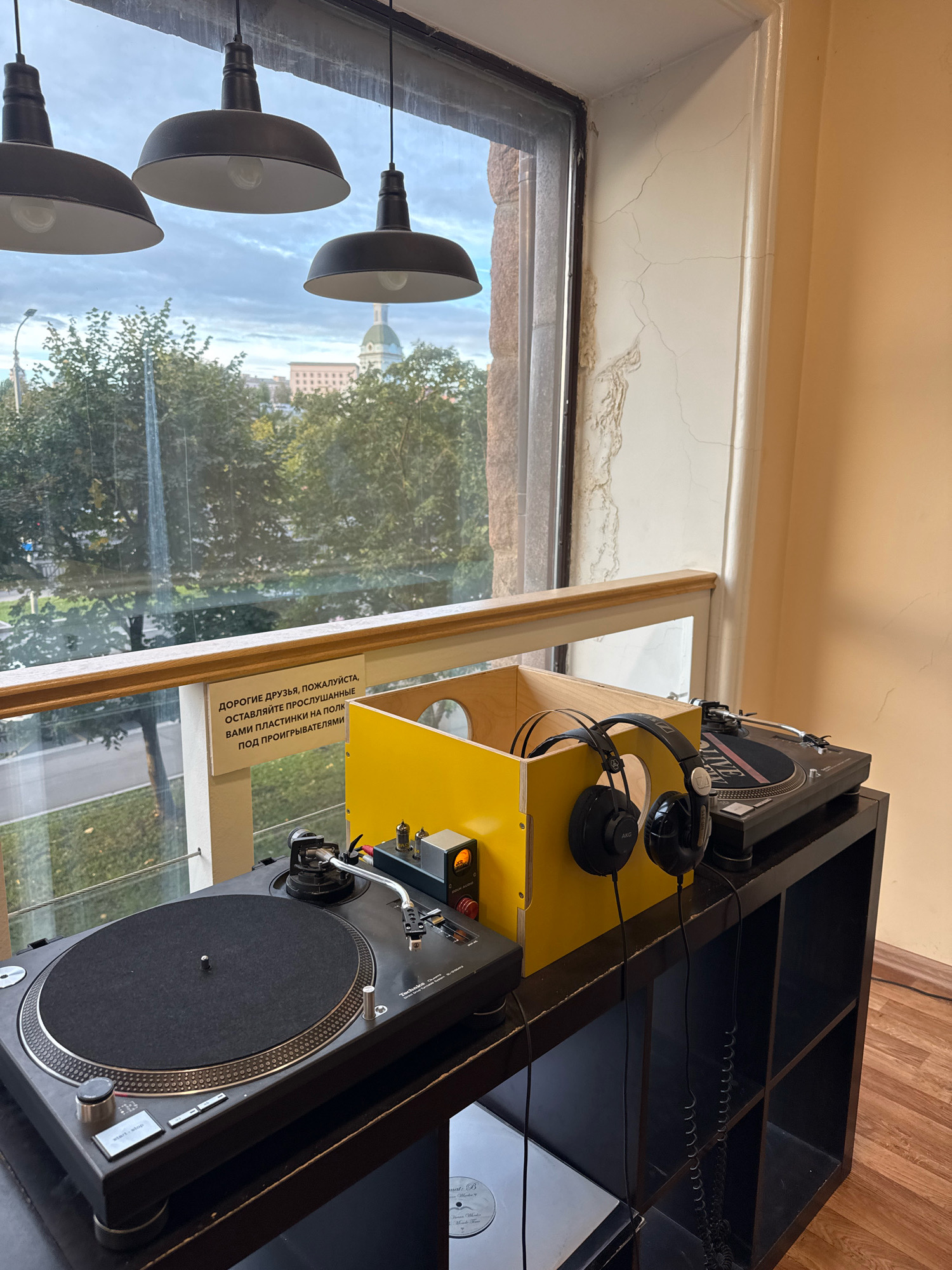 The image depicts a DJ setup positioned near a large window, overlooking a cityscape that includes trees, buildings, and a domed structure. The DJ setup consists of two turntables, headphones, and a small amplifier, all placed on a black shelving unit. A yellow box is also part of the setup, possibly for storage or as a visual element. A sign with Russian text is attached to a wooden railing in front of the window. Three pendant lights hang from the ceiling above the DJ equipment. The environment appears to be indoors, with a cream-colored wall visible to the right of the window. The view outside the window suggests an overcast day. The presence of the domed structure in the background suggests a possible location in Moscow, Russia, though this cannot be definitively confirmed from the image alone. There are no people visible in the image.
