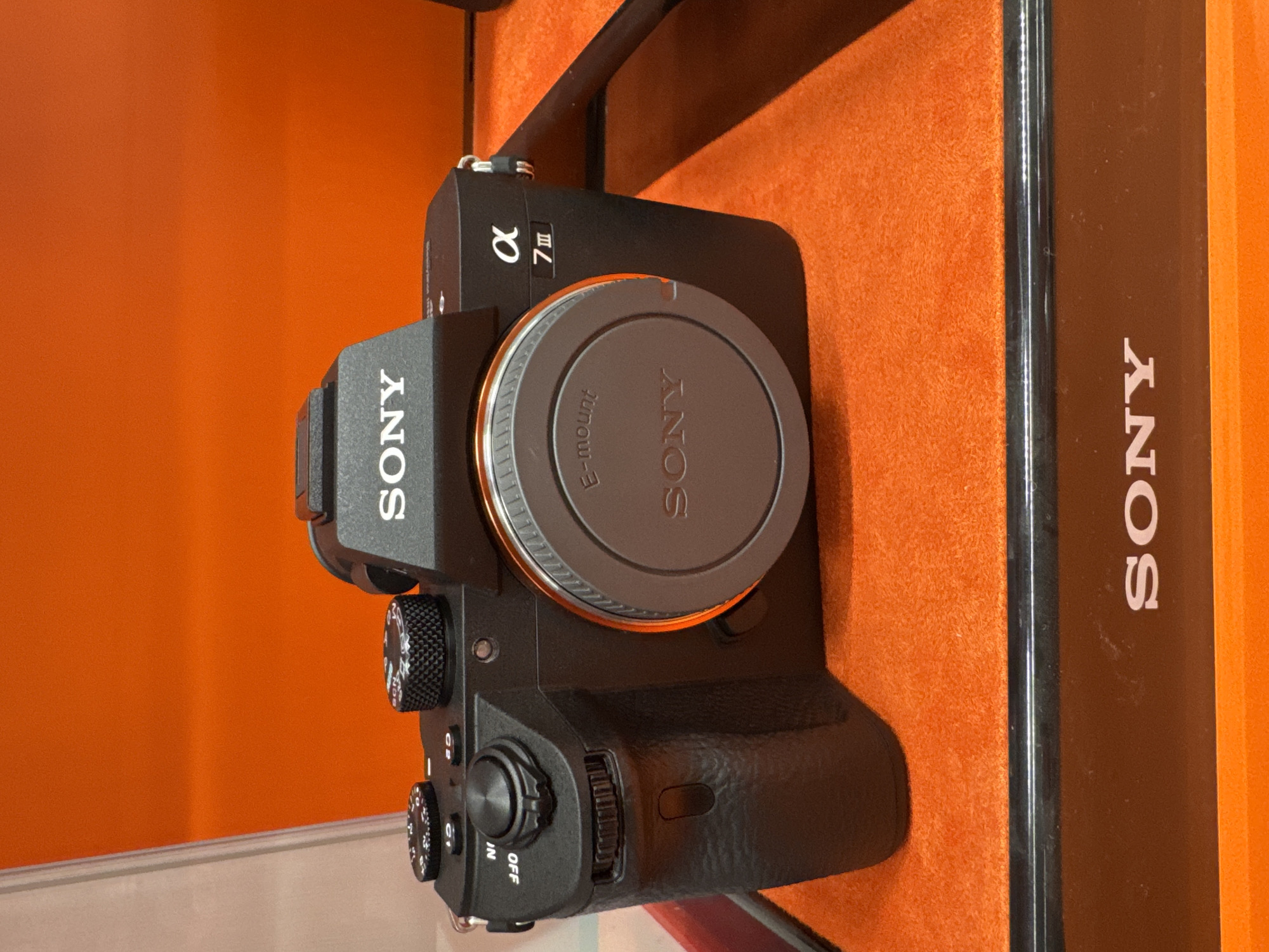 The image shows a Sony Alpha 7 III camera on display. The camera is black and has an E-mount lens cap. The camera and packaging is on an orange display shelf or backdrop. The packaging of the camera to the right has the brand name "SONY" on it. There are no people visible in the image. The location is not identifiable as being in Sofia, Bulgaria, from the provided image.