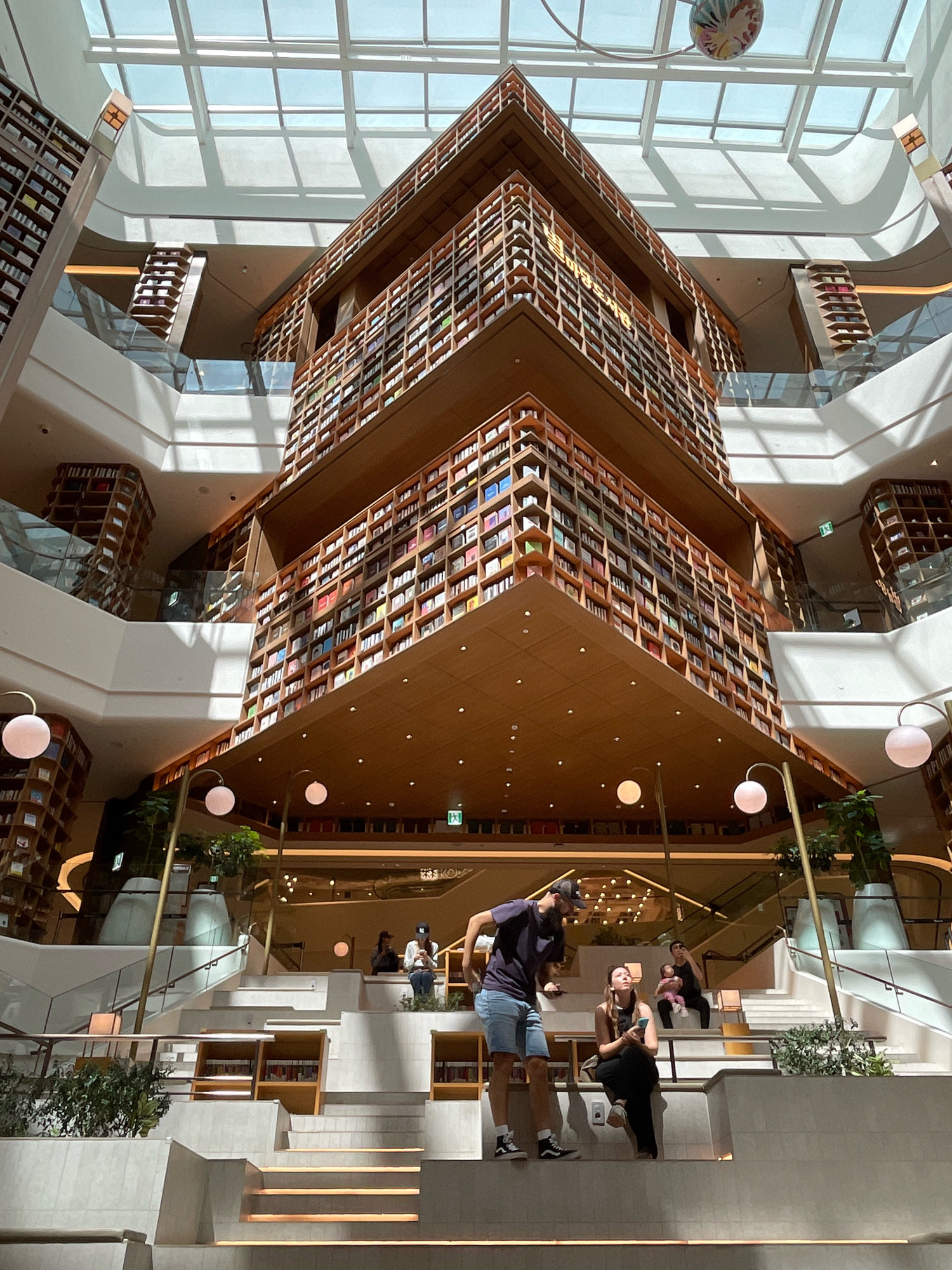 The image depicts the interior of Starfield Library, located in the Starfield Hanam mall in Hanam-si, Gyeonggi-do, South Korea. The library is characterized by its multi-story design featuring towering bookshelves filled with books that create a visually striking architectural element. Natural light streams in from a large glass ceiling, illuminating the space and casting shadows on the white staircases and platforms.
In the foreground, several individuals are present. A man wearing a dark t-shirt and denim shorts stands on a step, looking to his left. To his right, a woman in black attire is seated on a step, holding a smartphone. Behind her, another woman is seated with a baby in her lap, and further back, a person in a white shirt and a baseball cap is also seated. The people appear to be casually interacting or resting in the library space.
The environment is clean and modern, with a mix of wood and white architectural finishes. Various plants are integrated into the design, adding a touch of nature to the indoor setting. The overall impression is one of a spacious and well-lit public area designed for reading and relaxation, with a strong emphasis on the visual impact of the extensive book collections.