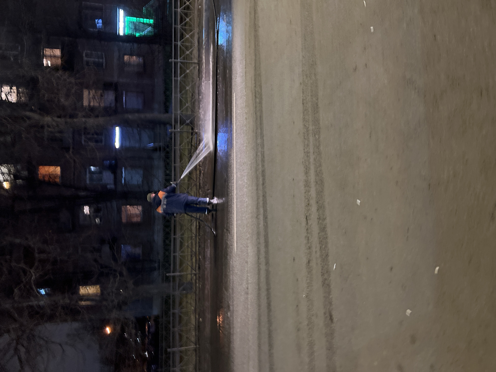 The image depicts a worker cleaning a wall or structure at night in what is likely Saint Petersburg, Russia. The worker, wearing dark clothing and a hat, is using a high-pressure water hose to spray a wall or tall structure. The wall is tall and potentially part of a building or fence. Behind the worker and the wall is a multi-story building with illuminated windows. In the foreground is a wet, paved surface, possibly a sidewalk or street. The lighting is dim, with illumination primarily from the building windows and the worker's spray.