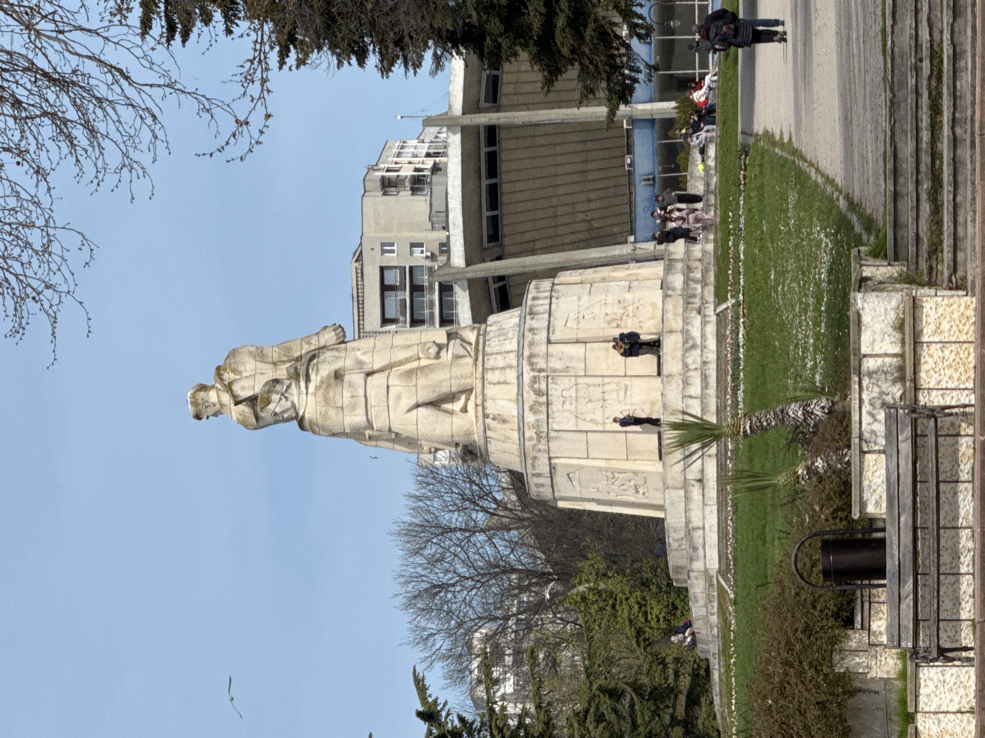 The image shows the Monument of the Soviet Army in Varna, Bulgaria, from a ground-level perspective. The monument is a large, light-colored stone structure topped with a statue of a soldier. A crowd of people are on the stairs of the monument. To the right of the monument, there is a landscaped area with grass, flowers, palm trees, and bushes. In the foreground, a bench and a trash can are visible. In the background, there are several buildings with a concrete façade. The sky is clear and blue.