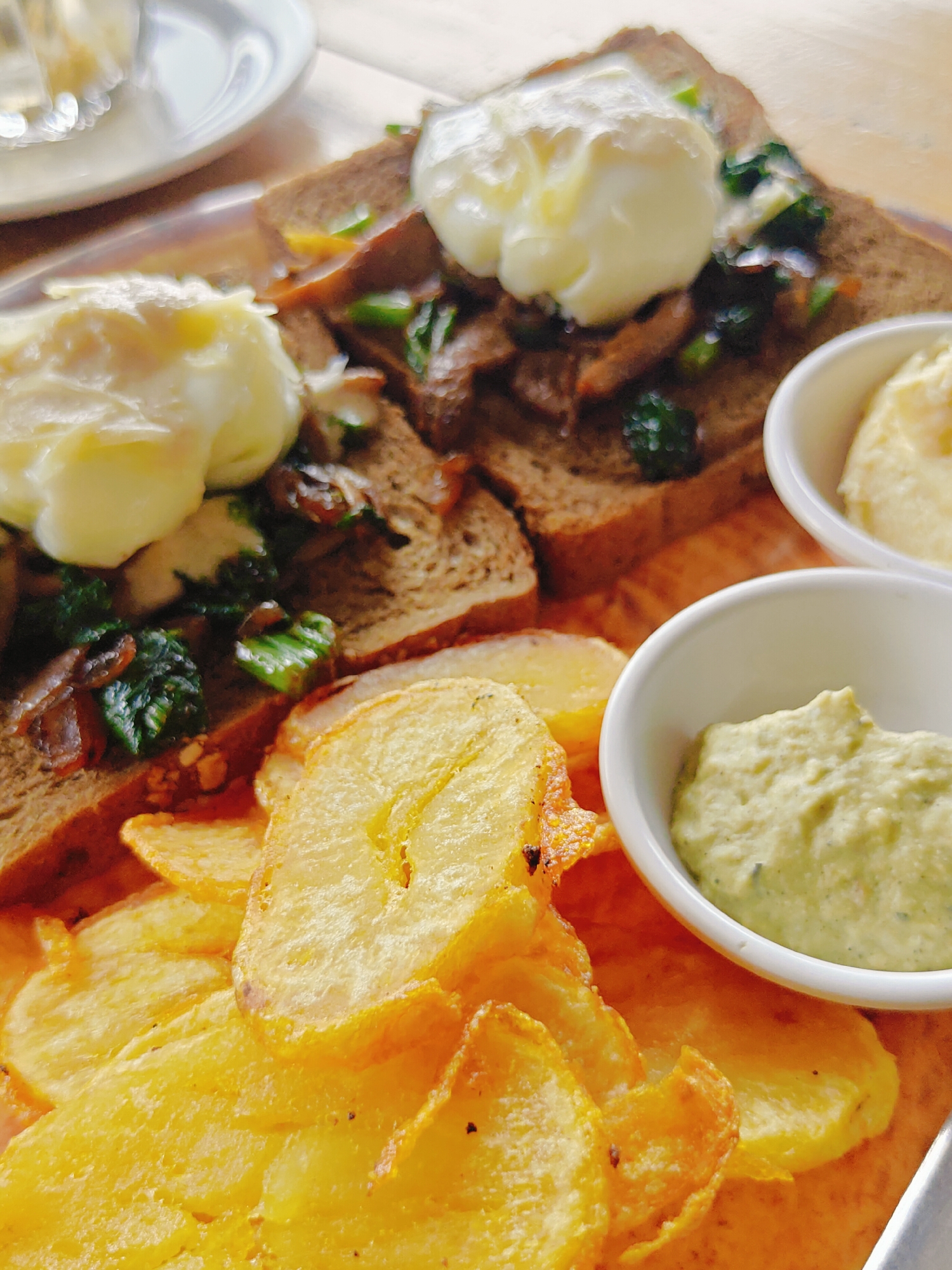 The image shows a close-up, overhead view of a breakfast dish served on a metallic platter. The dish consists of two slices of toasted bread, each topped with a poached egg and a mixture of greens and mushrooms. To the side of the bread, there are two small white bowls. One bowl contains a pale yellow, creamy substance, possibly mashed potatoes or hollandaise sauce. The other bowl contains a greenish, textured spread, likely an avocado-based dip. A pile of golden-brown potato chips is arranged in the foreground. The lighting suggests it is daytime, and the background is slightly blurred, hinting at an outdoor dining setting. This scene is likely from a cafe or restaurant in Pokhara, Nepal, known for its scenic beauty and diverse culinary offerings, including hearty breakfast options.