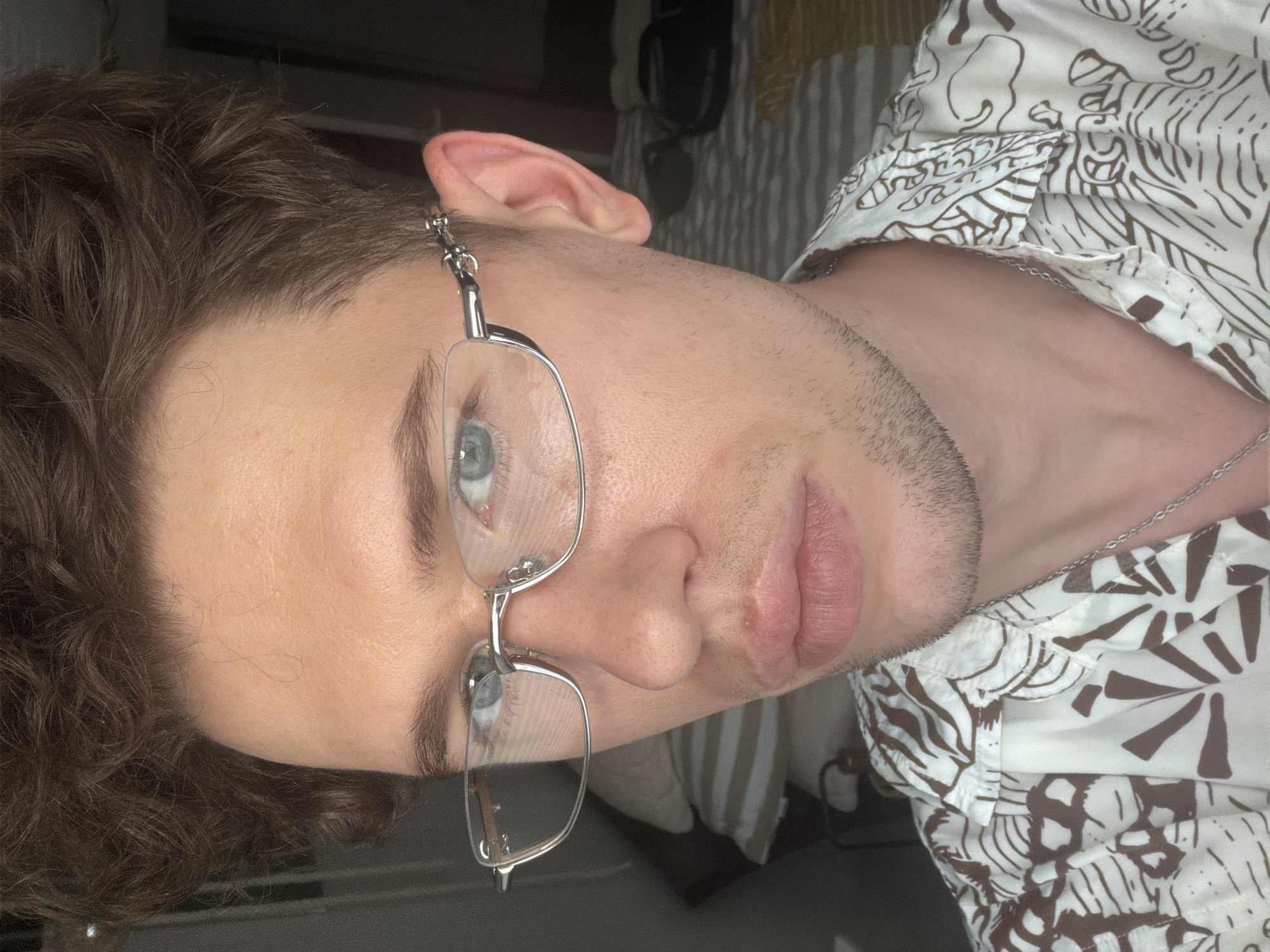 A close-up, head-and-shoulders shot captures a person with light skin, brown curly hair, and blue eyes. They are wearing thin-rimmed silver glasses. The person is wearing a white button-up shirt with a brown, abstract leaf-like pattern. A silver chain is visible around their neck. The background is blurred but appears to be an indoor setting with striped bedding and a dark, out-of-focus area to the right. There is no discernible indication of the city of Herne, Germany.