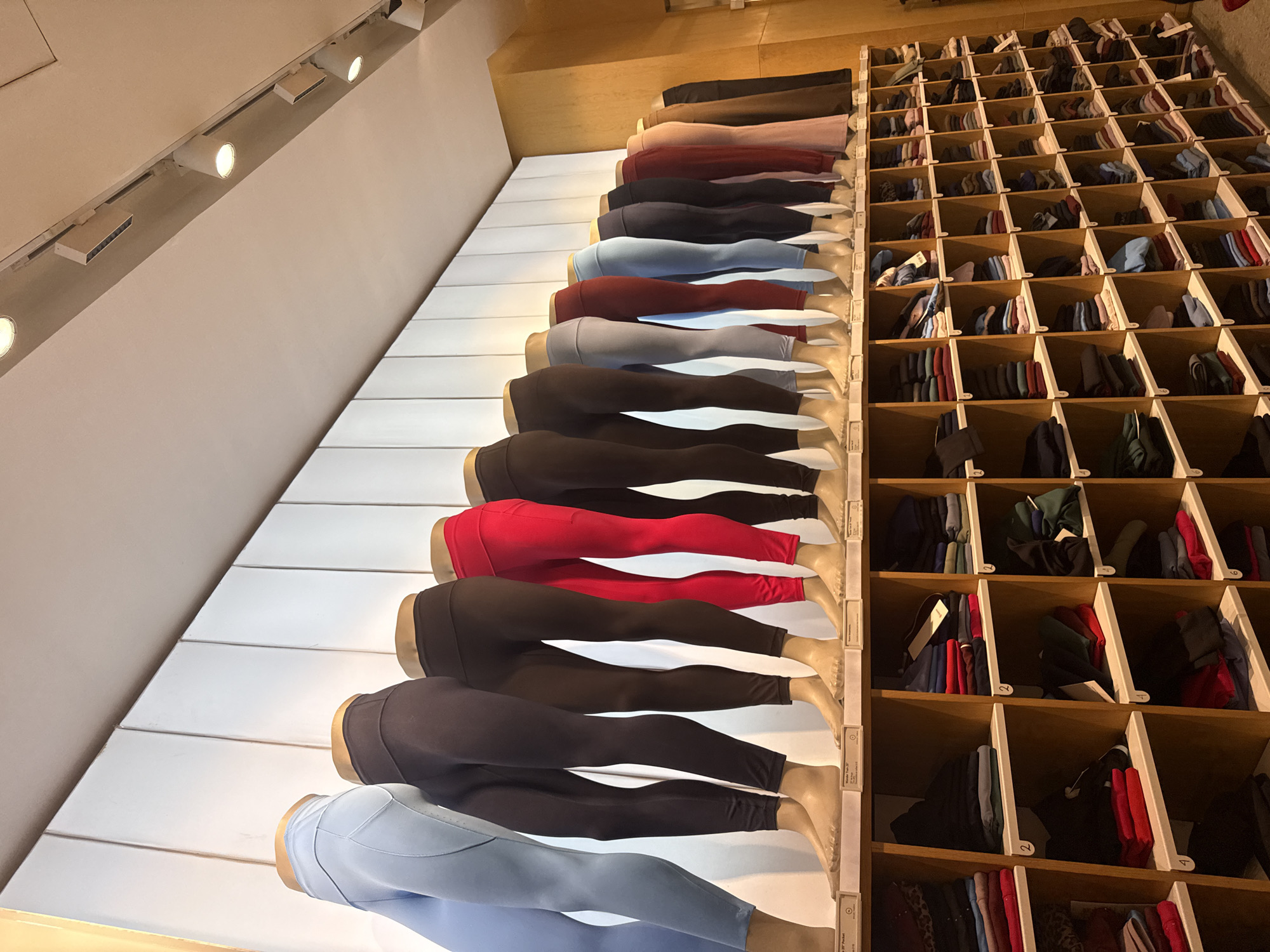 The image shows the interior of a retail store, likely a clothing store in Puebla, Mexico. Mannequin legs are arranged in a stepped display wearing different colored leggings. Next to the mannequins is a wall of stacked wooden cubbies filled with neatly folded clothing, possibly the same leggings in different colors and sizes. The lighting is bright and appears to be from track lights mounted on the ceiling.
