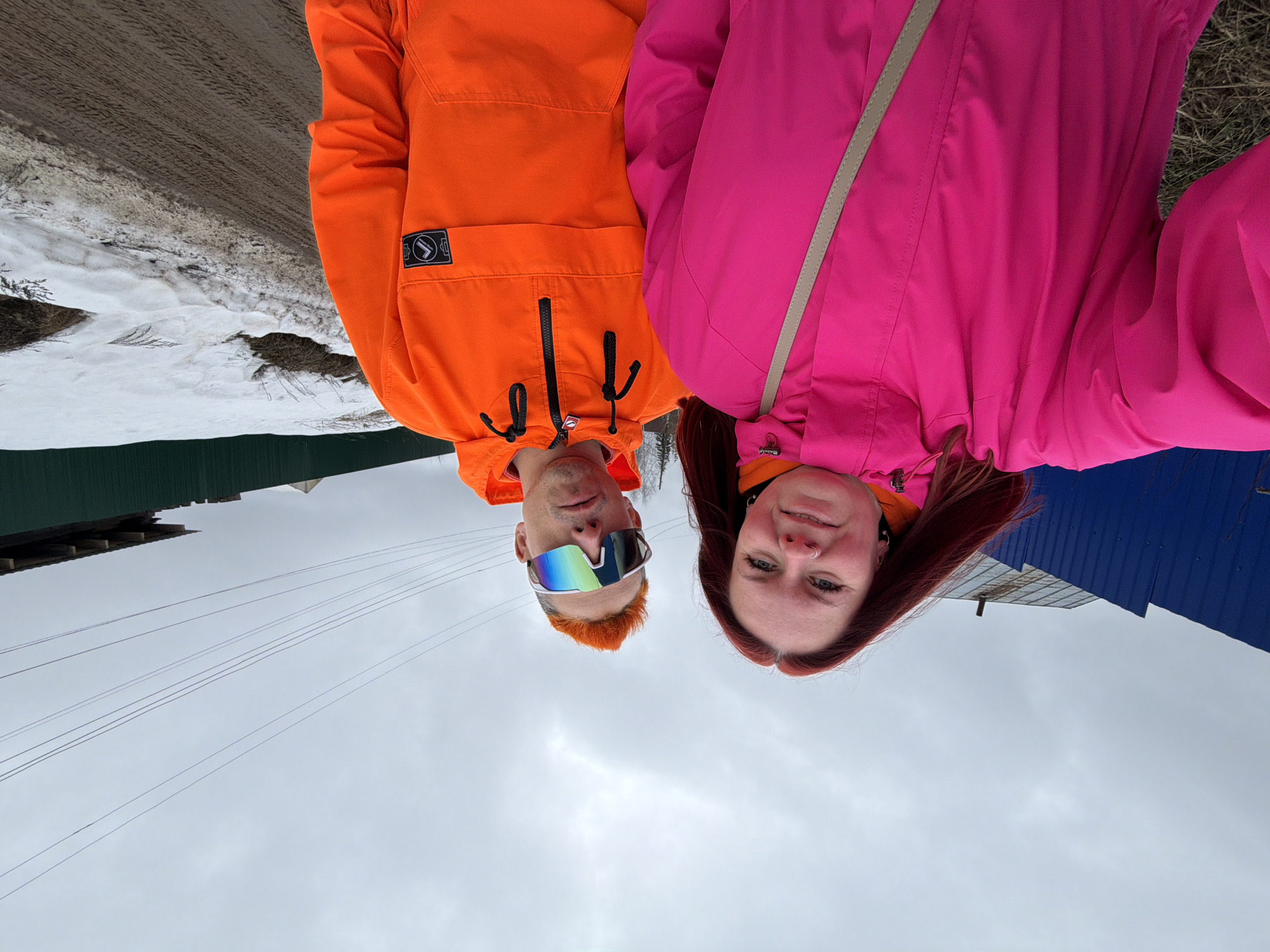 Two individuals are posing for a selfie in Chaikovsky, Russia. The man on the left wears orange outerwear and reflective sunglasses. The woman on the right wears pink outerwear and has auburn hair. The backdrop includes a cloudy sky, power lines, and residential structures with green and blue roofing. Patches of snow are visible in the background.