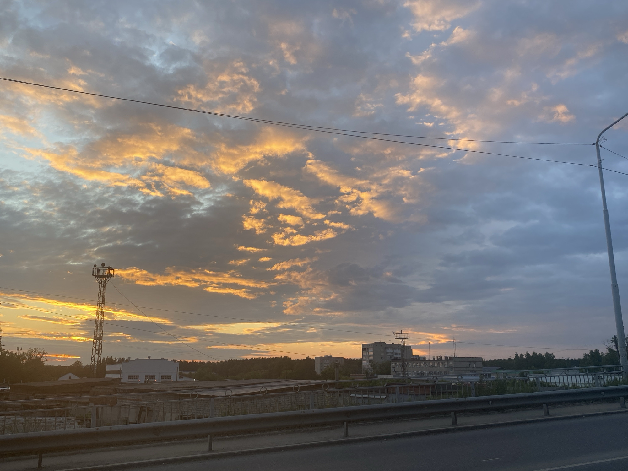 An outdoor, eye-level, long shot captures a wide expanse of sky filled with dramatic clouds tinged with orange and yellow hues from a sunset. The clouds are varied in texture, with some appearing wispy and others more dense. Two thin, black electrical wires stretch horizontally across the frame, dividing the sky. In the lower half of the image, the horizon features a line of trees and several buildings, some appearing to be industrial or commercial structures with flat roofs. A tall, lattice-like tower with lights on top stands prominently on the left side. A modern street light pole is visible on the far right. A road and a railing are in the foreground, suggesting the photo was taken from a bridge or elevated vantage point. The overall scene depicts a transitional period between day and night in what appears to be an urban or semi-urban setting.