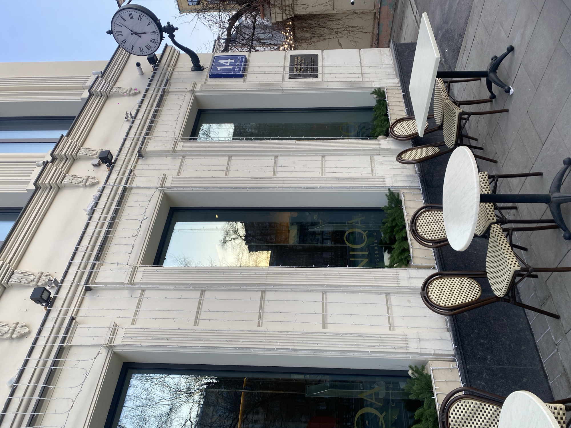 The image displays the exterior of a building housing UNIQA Patisserie & Cafe, located at Tverskaya Ulitsa, 14, Stroenie 5, Moscow, Russia. The light-colored building facade features three large ground-floor windows, flanked by vertical fluted pilasters and topped by decorative corbels. Cascading white string lights are suspended across the facade. Window boxes below the windows contain green fir branches. A blue rectangular street sign displaying "14 СТРОЕНИЕ 5" is mounted on the wall. To the right of this sign, a large, round clock with black Roman numerals (I-XII) and black hands indicates a time of approximately 1:55. A dark rectangular plaque with white Russian text is affixed below the blue sign. On the paved sidewalk, three unoccupied outdoor cafe tables are present: two round tables with light-colored tops and dark bases, and one rectangular table with a similar light-colored top and a dark, four-pronged base. Five bistro-style chairs with dark brown frames and woven light/dark patterned seats and backrests are arranged around the tables. The windows reflect bare tree branches, parts of distant buildings, and a clear sky. No individuals are visible.