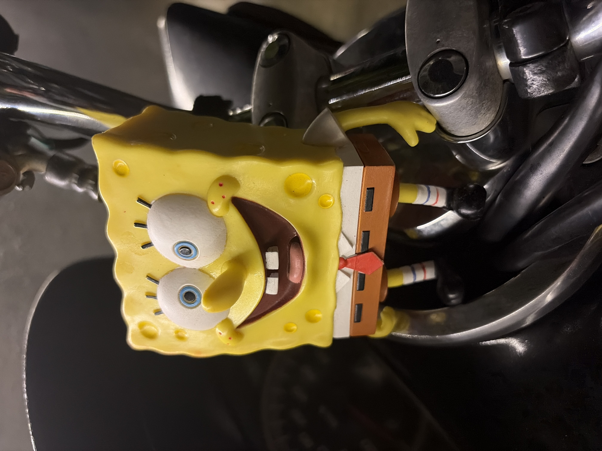 A Spongebob Squarepants figurine is mounted on the handlebars of a motorcycle. The figurine is bright yellow, with blue eyes, a wide smile showing square teeth and pink gums, and red cheeks. It wears a white shirt, a red tie, and brown pants with a white belt. The motorcycle handlebars are made of black and chrome metal. The background is dark and out of focus, suggesting an indoor or nighttime setting. The scene is a close-up shot, focusing on the Spongebob figurine and its immediate surroundings on the motorcycle.
