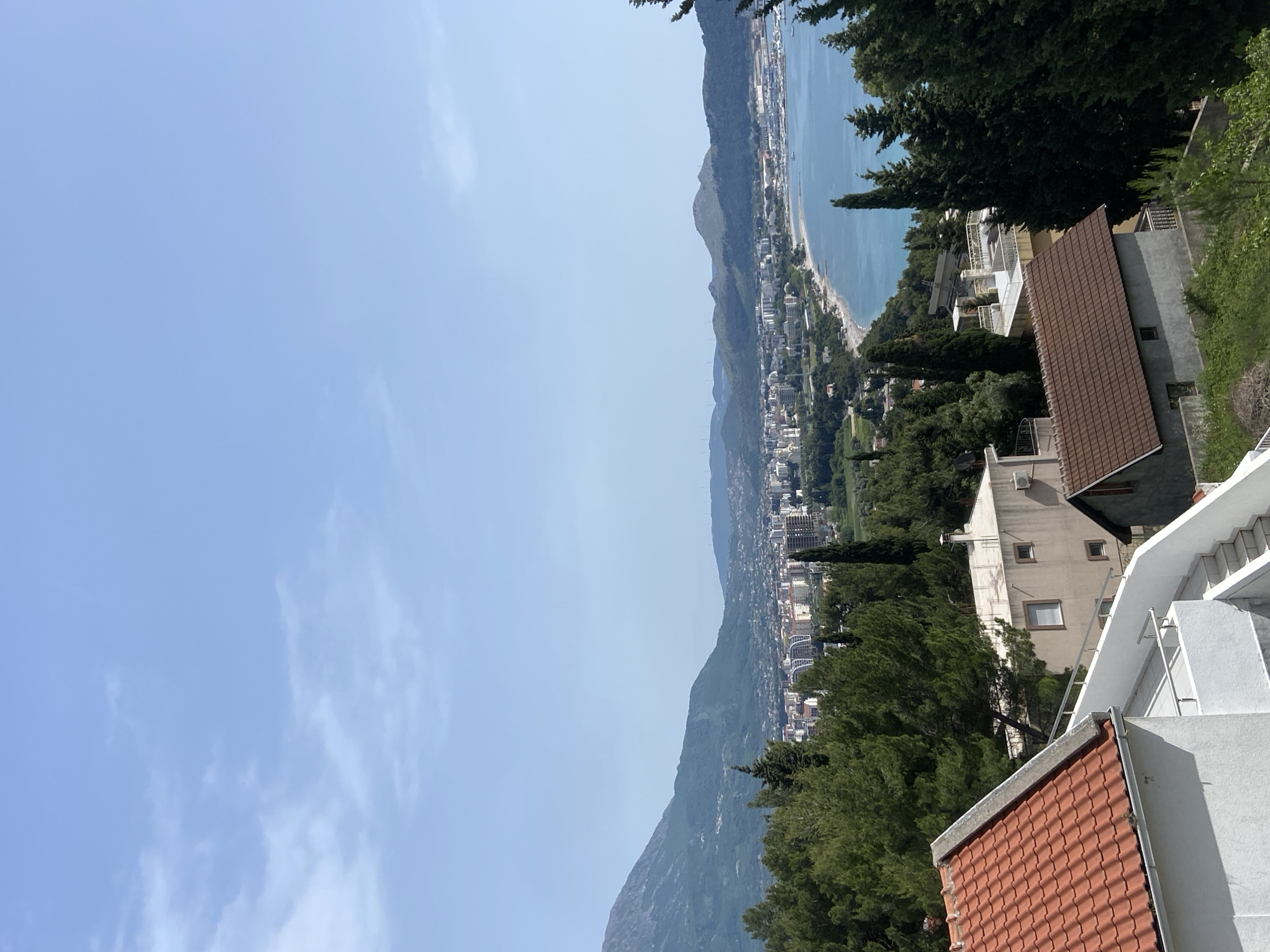 A high-angle, eye-level photograph shows a view of Šušanj, Montenegro. The sky is blue with wispy clouds. The Adriatic Sea stretches along the shoreline, with a beach visible. Mountains rise parallel to the coast, partially obscured by haze. The cityscape includes buildings, trees, and vegetation. Foreground details feature rooftops, trees, and partial views of residential buildings. The photograph is taken from an elevated position, possibly a hillside or balcony.