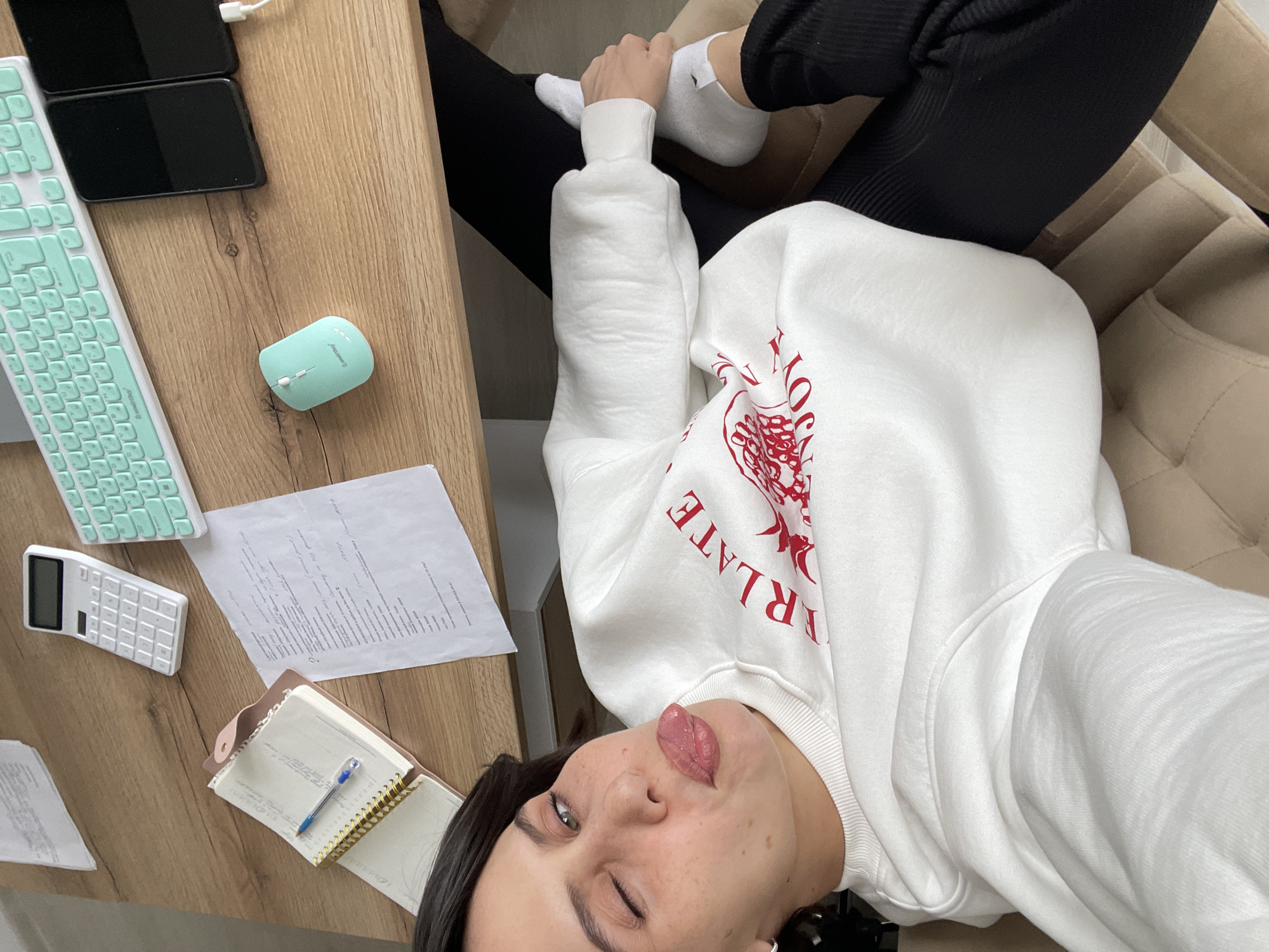 A person in a white sweatshirt and black pants is lying in a beige armchair, with their feet up on a wooden desk. The desk has a turquoise keyboard and mouse, two black smartphones, a white calculator, a notebook with a pen, and a sheet of paper with text. The person is sticking their tongue out. The image does not provide enough information to determine the exact location in Izhevsk, Russia.