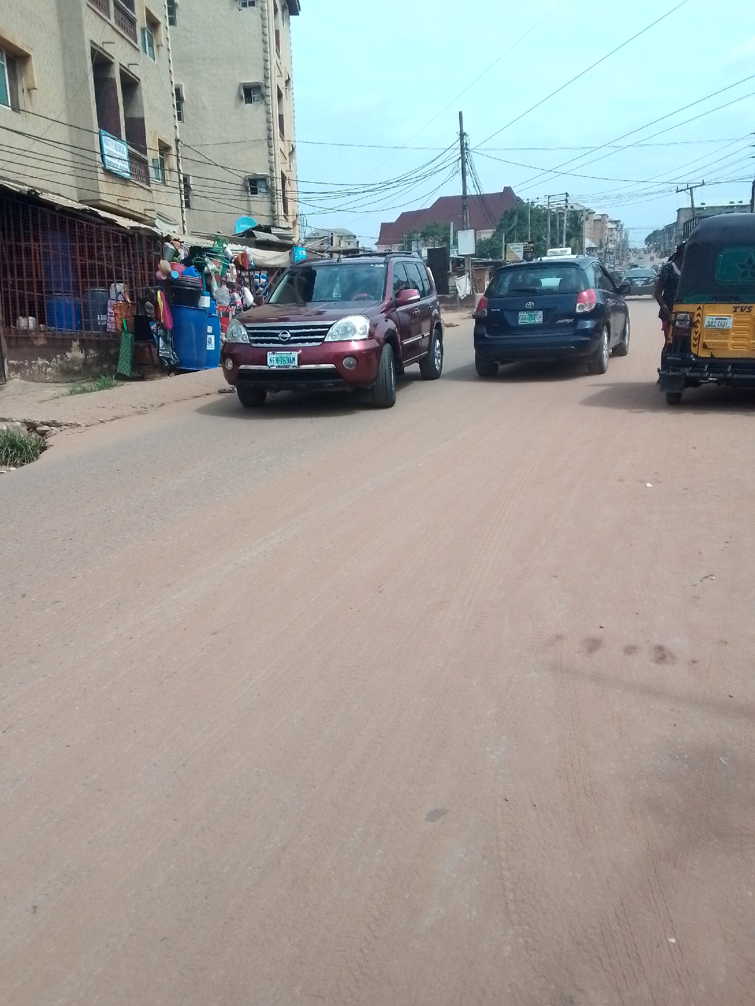 This is a street-level view of a road in Nkpor, Nigeria, likely on a moderately overcast day. In the foreground, the reddish-brown, dusty road shows tire tracks. On the left side of the road, a light-colored, multi-story building with balconies and several windows stands adjacent to a market area. This market features a metal fence, various goods displayed for sale, including blue barrels and other containers. A maroon SUV with a license plate reading "NEN 763 AM" is parked facing away from the viewer. Further down the street, a dark blue car with a license plate "GGE 702GH" is also moving away. To the right, a yellow motorized tricycle, commonly known as a Keke, with a license plate "UMZ 2E YI" is visible, also moving away. Several utility poles and electrical wires are prominent throughout the scene, indicating the presence of infrastructure. The overall impression is a busy, urban street scene typical of many Nigerian cities.
