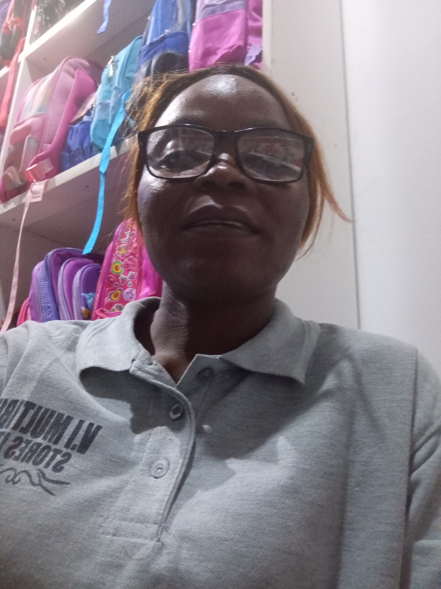 A mid-shot, medium-light skin-toned woman with brown hair and eyeglasses is positioned in the center of the frame, looking directly at the camera. She is wearing a grey polo shirt. Behind her, on shelves, are several backpacks of various colors, including pink, blue, and purple. The background appears to be an indoor setting, possibly a room with a white wall to the right. The scene is well-lit, with some reflections visible in the woman's eyeglasses. The image is of moderate resolution and appears to have been taken with a mobile device. The scene is not identifiable as being in Howayi, Nigeria, or any other specific location.