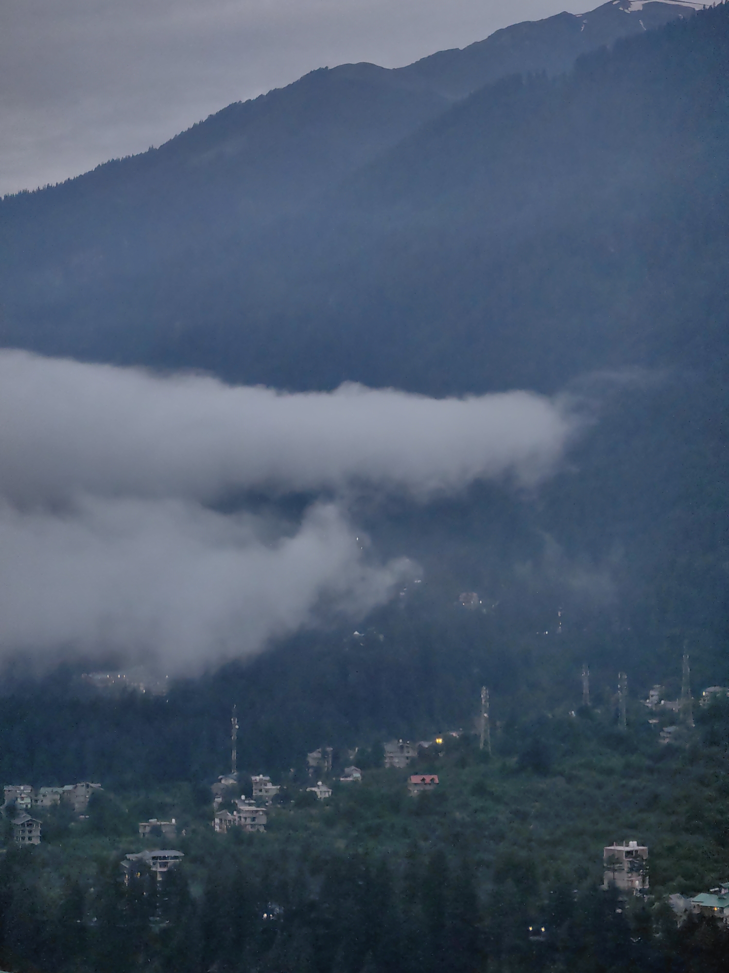 This is a landscape photograph taken in Vashisht, India, during twilight. The image depicts a mountain village nestled in a valley, with several buildings scattered across the slopes. The dominant feature of the image is the imposing mountain range that occupies the upper portion of the frame, shrouded in a layer of atmospheric mist and fog. The mountains are dark blue and green, with the very top of the highest peak showing a hint of snow. A thick band of white cloud hangs low across the mountainside, partially obscuring the village below. The village itself is characterized by a cluster of buildings, many of which appear to be multi-story residential structures. Several telecommunication towers are visible, standing tall amidst the greenery. The lighting suggests it is either early morning or late evening, with a subdued, diffused light bathing the scene. The overall atmosphere is serene and somewhat melancholic, with the mist and twilight creating a sense of depth and mystery.