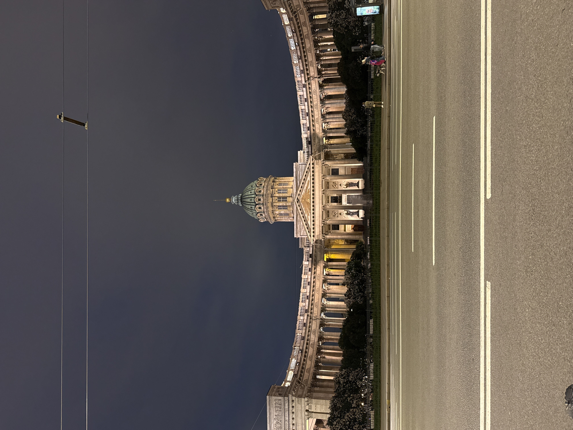The image captures the Kazan Cathedral in Saint Petersburg, Russia, at night. The grand neoclassical cathedral, with its semicircular colonnade and central dome, is illuminated, creating a striking contrast against the dark sky. In the foreground, a wide asphalt road with white lane markings stretches across the frame. Several people are visible on the sidewalk in front of the cathedral, including a person walking a dog. Trees line the area between the road and the building. Overhead, power lines are strung across the sky, one with a visible support structure. The overall scene depicts a well-lit architectural landmark in an urban setting at night.