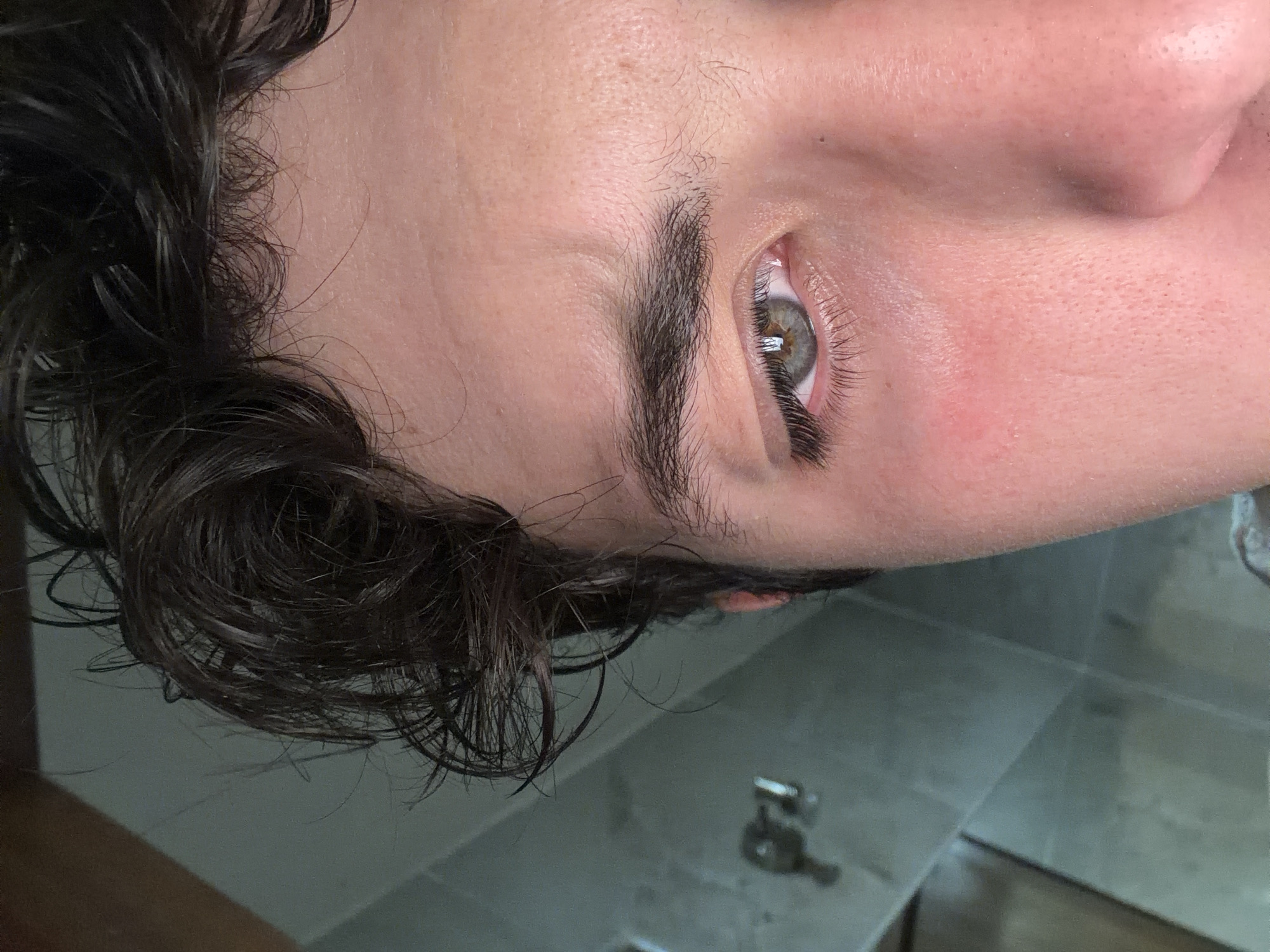 An image of the right side of a person's face, oriented so that their eye is in the center. The person has dark hair, a dark eyebrow, and green eyes. The skin around their eye is pink, and there are visible pores. The background is a tiled surface, possibly a bathroom. There are also some small objects visible on the tiled surface, but they are out of focus. The person's face is close to the camera and fills much of the frame. The setting cannot be verified as San Andrés Cholula, Mexico, from the image alone.