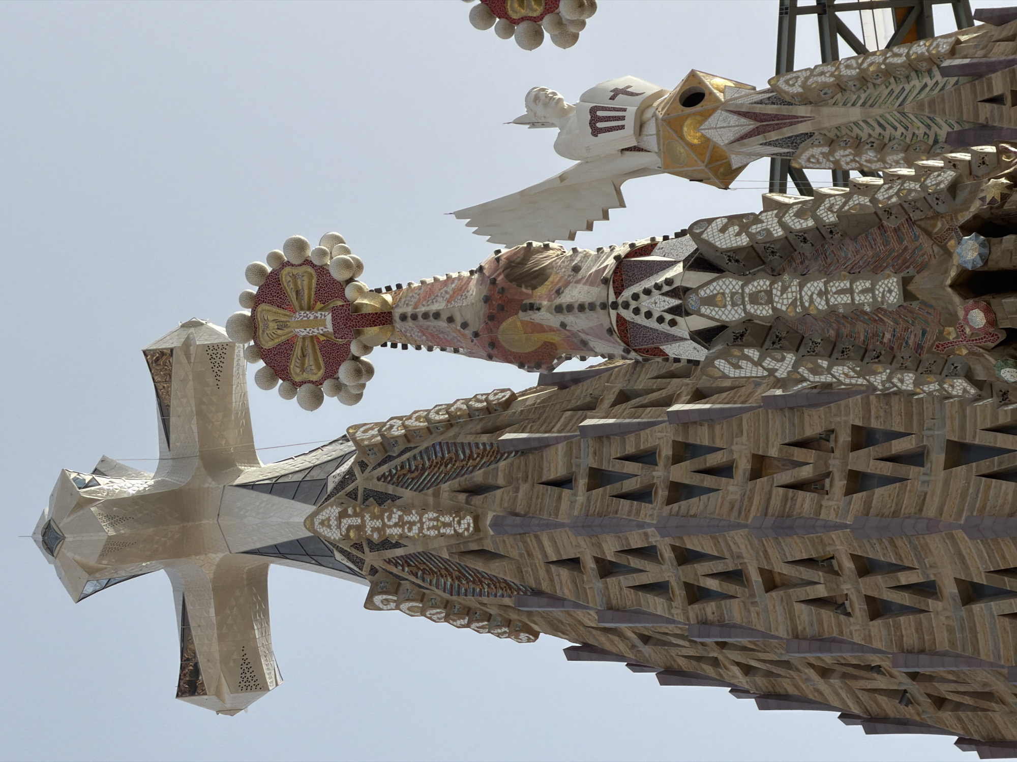 This is a low-angle shot of the spires of the Sagrada Familia, a basilica in Barcelona, Spain. The image is rotated 90 degrees clockwise, so the spires appear to be extending upwards from the bottom of the frame. The sky is a pale, clear blue, suggesting daytime with fair weather.
The spires are intricately decorated with mosaics and stone carvings. One spire features a white statue of a figure with outstretched arms and wings, possibly an angel, with a cross and a stylized letter "M" on its chest. Another spire is adorned with a large, circular mosaic featuring red, gold, and white patterns, surrounded by small, spherical white elements. Further down, the spires display geometric patterns in stone and mosaic, with triangular openings that add to their textured appearance. The overall style is characteristic of Antoni Gaudí's unique architectural approach, blending organic forms with religious symbolism.
The angle and detail suggest a close-up view of the upper portions of the basilica, focusing on the elaborate craftsmanship of its facade. The reflection of the spires in what appears to be a mirrored surface or water at the bottom of the frame adds a surreal and symmetrical element to the composition. No visible text is present in the image.