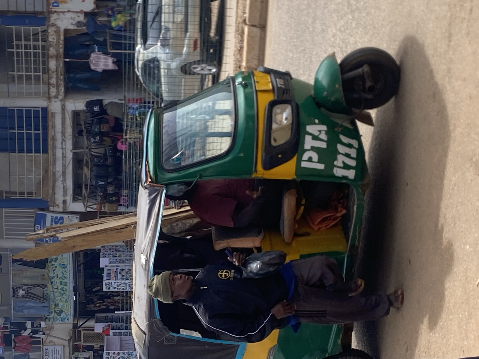 This image captures a street scene in a commercial area within Bukuru, Nigeria, featuring a three-wheeled vehicle and multiple individuals.
In the foreground, a green and yellow auto-rickshaw (locally known as a Keke Napep) is prominent. The vehicle's body is green with a yellow stripe, and "PTA 1711" is printed in white on its lower front. The front also displays a TVS brand logo. Several long wooden planks, appearing weathered, are secured on the black fabric roof of the tricycle, extending upwards.
A Black male, appearing to be a young adult, stands on the left side of the tricycle. He is wearing an olive green beanie, a dark blue track jacket with white stripes along the sleeves and a yellow circular emblem on the left chest (containing Chinese characters "忠義堂"), a blue undershirt, dark grey track pants with three white stripes down the sides, and brown sandals. His left foot is on the ground while his right foot is slightly raised, suggesting movement or a momentary stop. He holds a dark plastic bag in his left hand and a small, dark rectangular object in his right hand, looking towards the right side of the frame.
Inside the tricycle, a portion of another individual, presumably the driver, is visible, wearing a maroon or dark red short-sleeved shirt. Another dark-clad figure is partially visible further back in the passenger compartment.
The background consists of multi-story buildings housing various retail shops. The building on the left features displays of shoes, bags, and clothing items on the ground floor, with a sign partially visible that reads "ONE." The upper floor also displays clothing. The building to the right exhibits more clothing displays, including a pink plaid shirt and blue garments, along with bags and a cluster of green and yellow balloons near an entrance. A sign with an "M" logo ("M Point") is visible on the upper section of the left building. The architecture includes blue-gridded windows and white balcony railings on the upper floors.
Behind a metal barrier on the right side of the road, a silver SUV is parked. The ground is an asphalt surface, appearing dry and somewhat uneven. The scene suggests a bustling urban environment under daylight conditions.