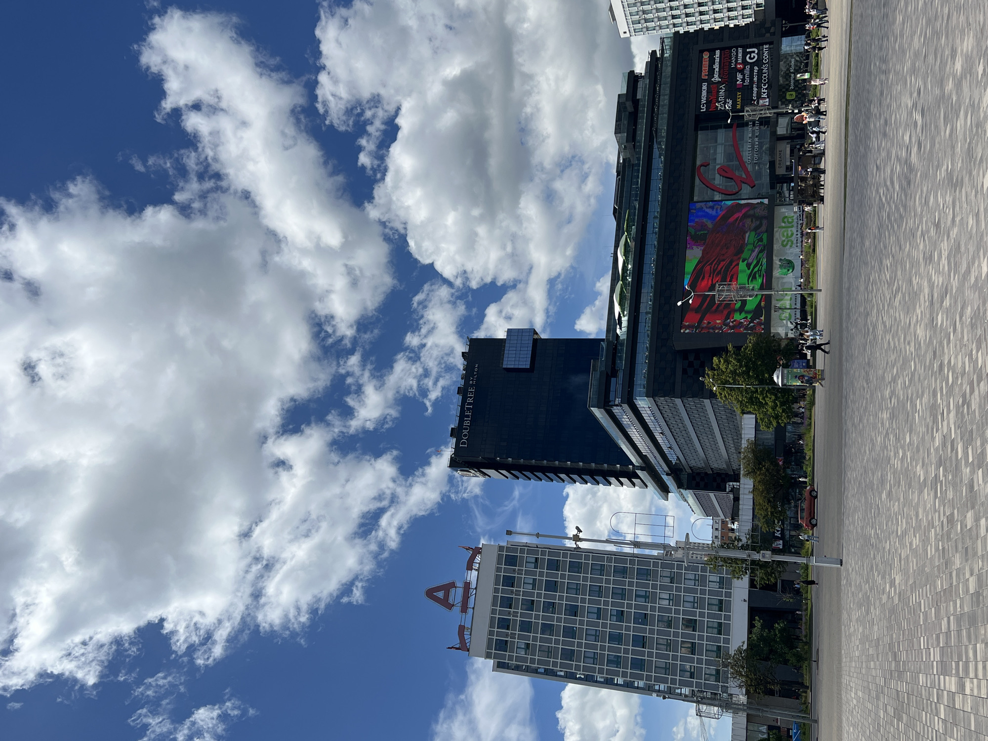 This media file shows an outdoor scene in Minsk, Belarus, featuring modern architecture and a bright, cloudy sky. In the foreground, a wide, paved plaza stretches out, with a pattern of rectangular stones. Several tall buildings dominate the background.
One prominent building is a dark, glass-fronted structure with the "DOUBLETREE HILTON" logo visible on its side. Adjacent to it is a larger, darker building adorned with various brand logos and digital screens. These include "LC WAIKIKI," "STRADIVARIUS," "ZARINA," "MANGO," "MF 5," "familia," "GJ," "KFC," and "COLIN'S." A large red "C" logo for "GALLERIA MINSK - ТОРГОВЫЙ ЦЕНТР" is also displayed. One of the screens shows a vibrant, colorful graphic with a figure in red.
Another building in the frame is a lighter-colored, grid-patterned tower with a large red "A" logo at its peak. Scattered throughout the scene are small groups of people, suggesting a public space or shopping area with moderate foot traffic. Trees are visible, indicating a temperate season. The sky is a vibrant blue, punctuated by large, fluffy white clouds, suggesting daytime with dynamic weather. The overall impression is one of a bustling urban environment on a pleasant, albeit cloudy, day.