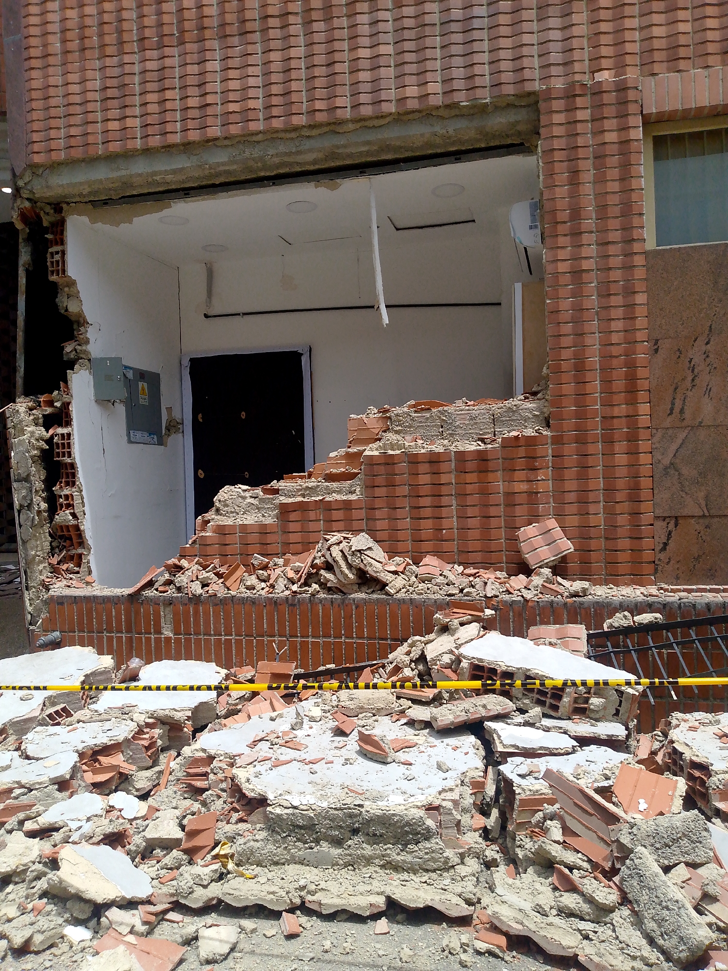 The image depicts the aftermath of a structural collapse on a building's facade, likely due to an earthquake or similar event. The scene is chaotic, with a large section of the brick exterior wall, including a doorway and part of the interior, destroyed. Rubble, consisting of bricks, concrete, and insulation, is scattered across the ground in front of the damaged area. Yellow caution tape with black stripes is stretched across some of the debris, indicating a hazardous zone.
The building itself is constructed with red bricks, featuring a distinctive textured pattern on the facade. An air conditioning unit is visible on the interior wall of the damaged section. A gray electrical panel with a warning symbol is mounted on the white interior wall to the left of the destroyed opening. A dark door is visible behind the debris.
The lighting suggests it is daytime, with no clear indication of weather conditions. The overall impression is one of destruction and a recent catastrophic event. The location is identified as Caracas, Venezuela, but no specific landmarks are visible in the image. There is no visible text beyond the warning symbol on the electrical panel and the caution tape.