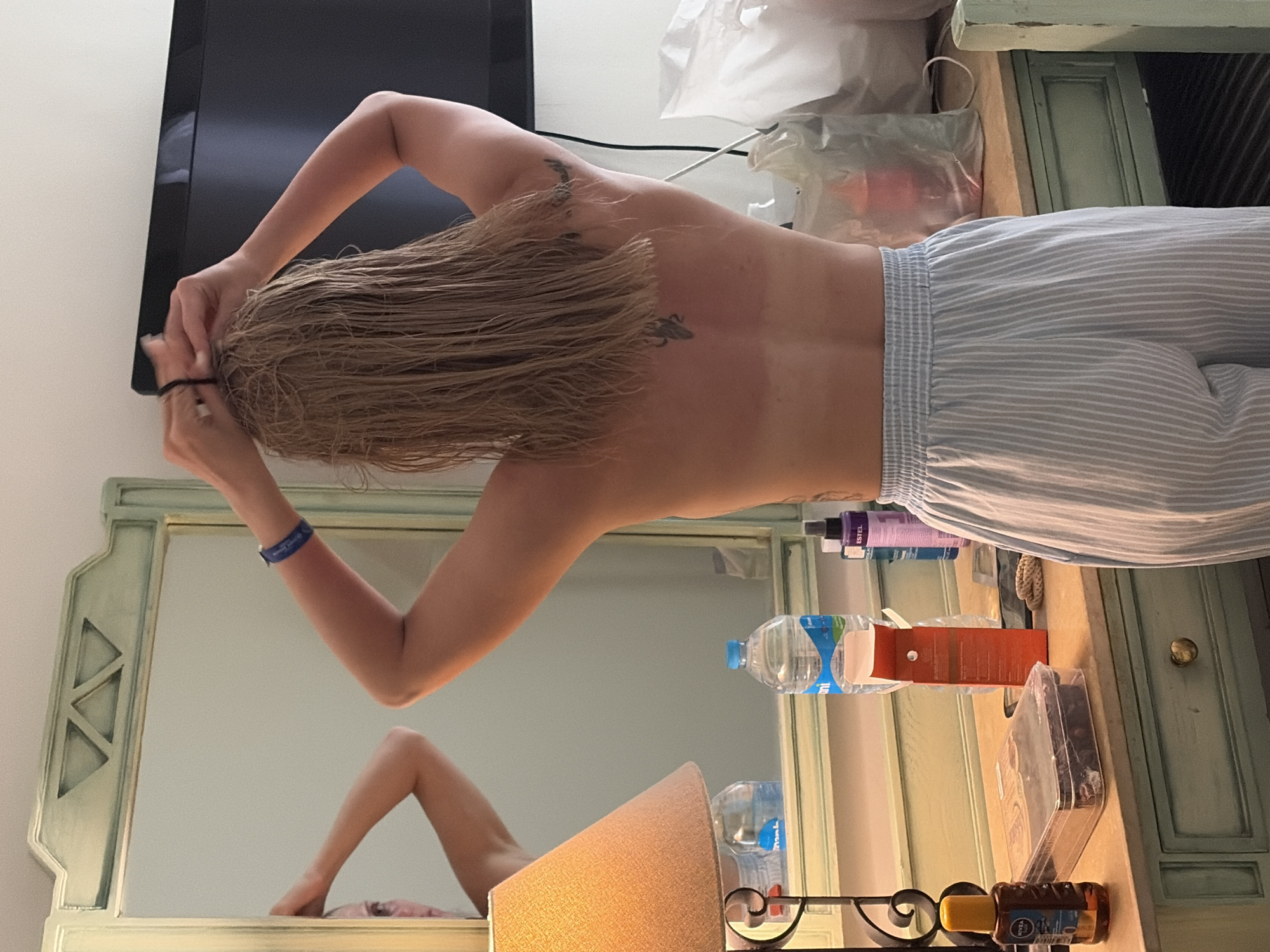 The image depicts a person with wet, blonde hair, tying it up with a hair tie. They are wearing striped pajamas and have tattoos on their upper back and arm. The person is standing in front of a dressing table with a mirror that reflects their arm and a portion of their face. The dressing table has several items on it, including a water bottle, a brown box, beauty products, and a lamp. A television is mounted on the wall behind the dressing table. The location is likely a hotel room in Sharm El Sheikh, Egypt, based on the interior design and items present in the room.