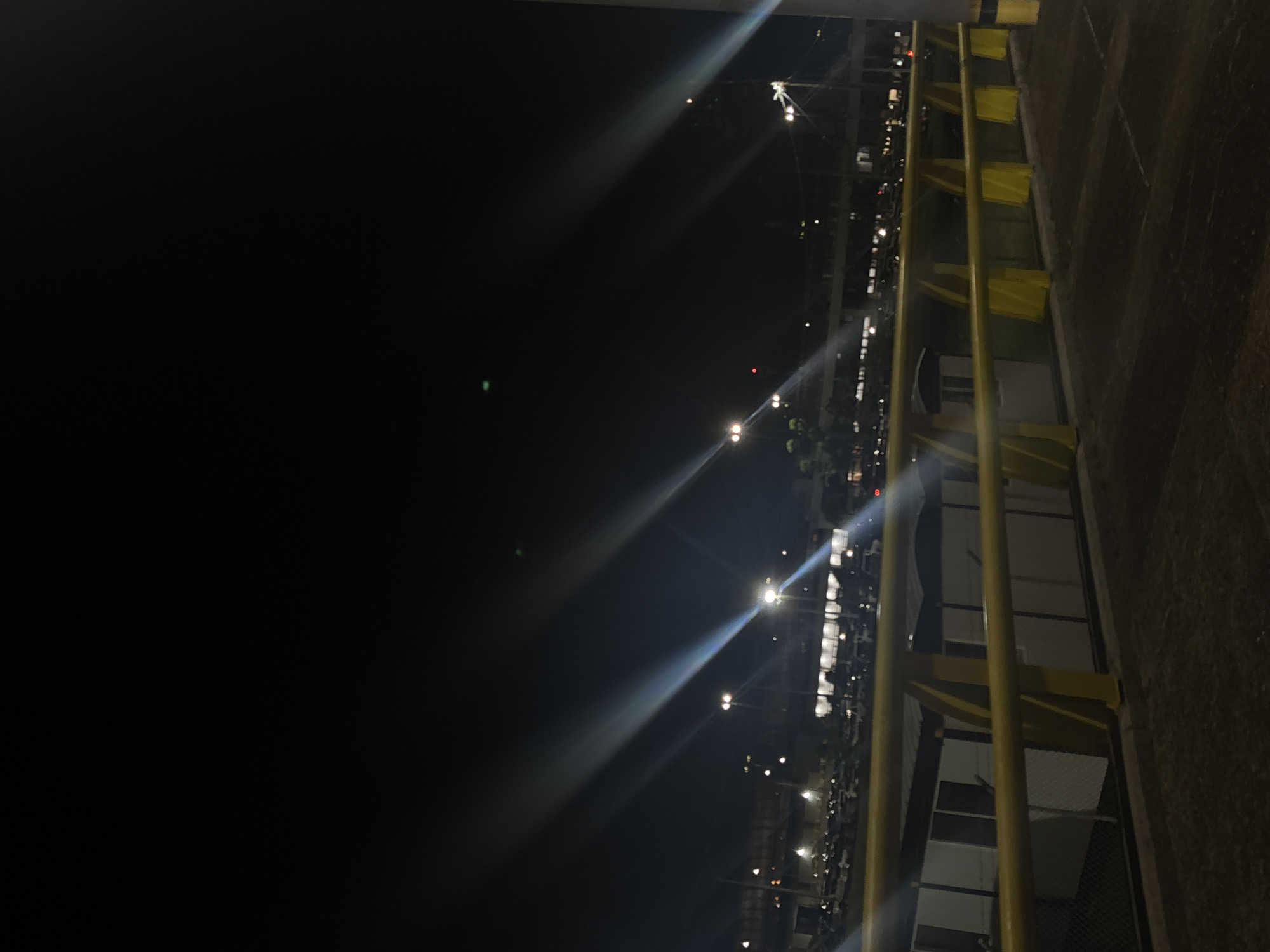 At night in Rionegro, Colombia, a parking lot illuminated by streetlights is seen from a slightly elevated position. Yellow safety railings run along the right side of the frame, partially obscuring a building with visible windows and mesh panels. Beyond the railings, rows of parked cars stretch into the distance, lit by more streetlights. The sky above is dark, with light flares around the streetlights.