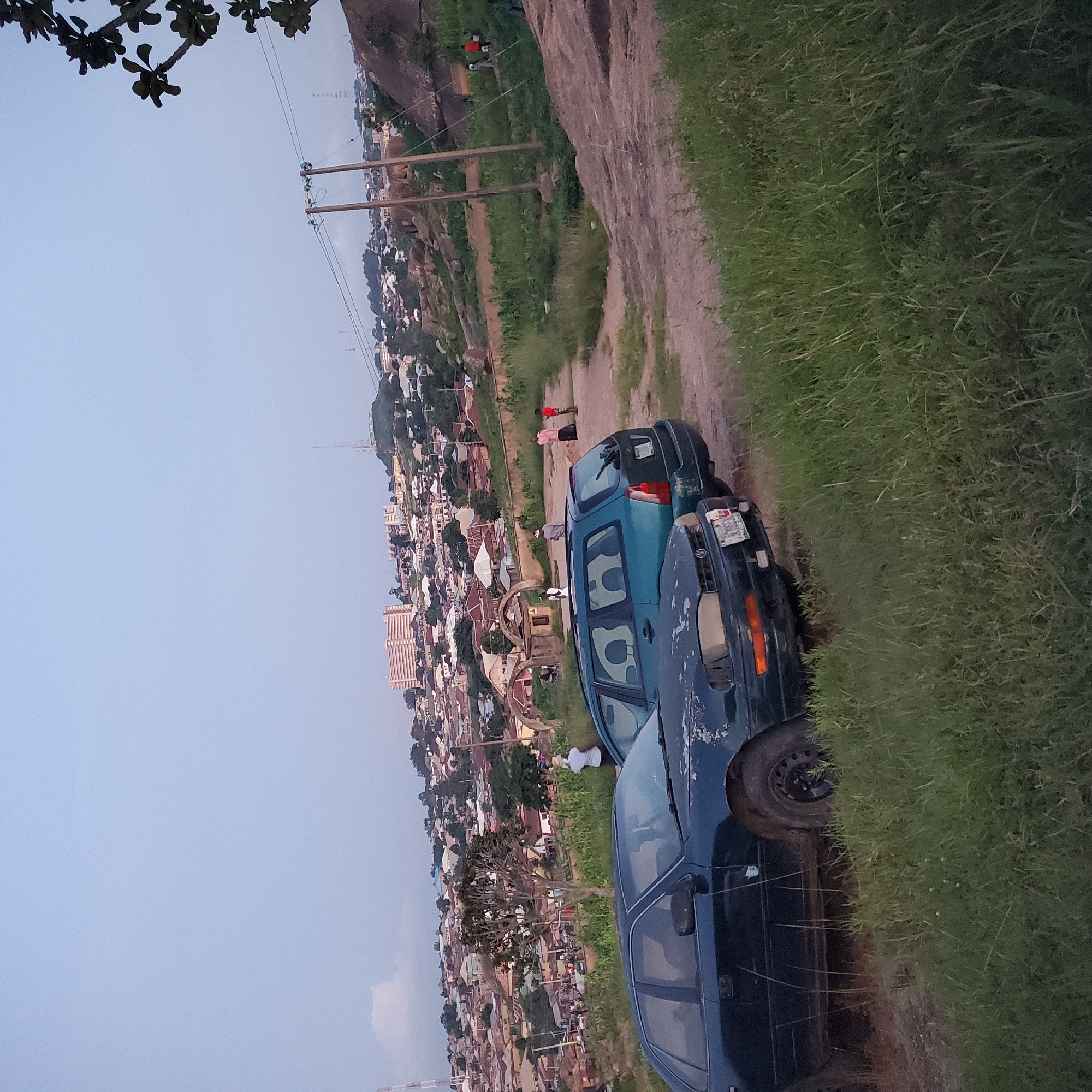 The image captures a landscape scene in the city of Jos, Nigeria, characterized by a vast expanse of grassy terrain dotted with rocks and a sprawling urban settlement in the background. Two cars are parked in the foreground, a dark sedan and a teal station wagon, both appearing weathered and in need of maintenance. Several people are visible in the middle ground and background, some appearing to be engaged in conversation or walking. The settlement in the background is densely populated with a mix of residential buildings and some larger structures, including a prominent multi-story building that may be a hotel or apartment complex. The overall impression is one of a rural or semi-rural area adjacent to a developed urban center, under a clear, light blue sky.