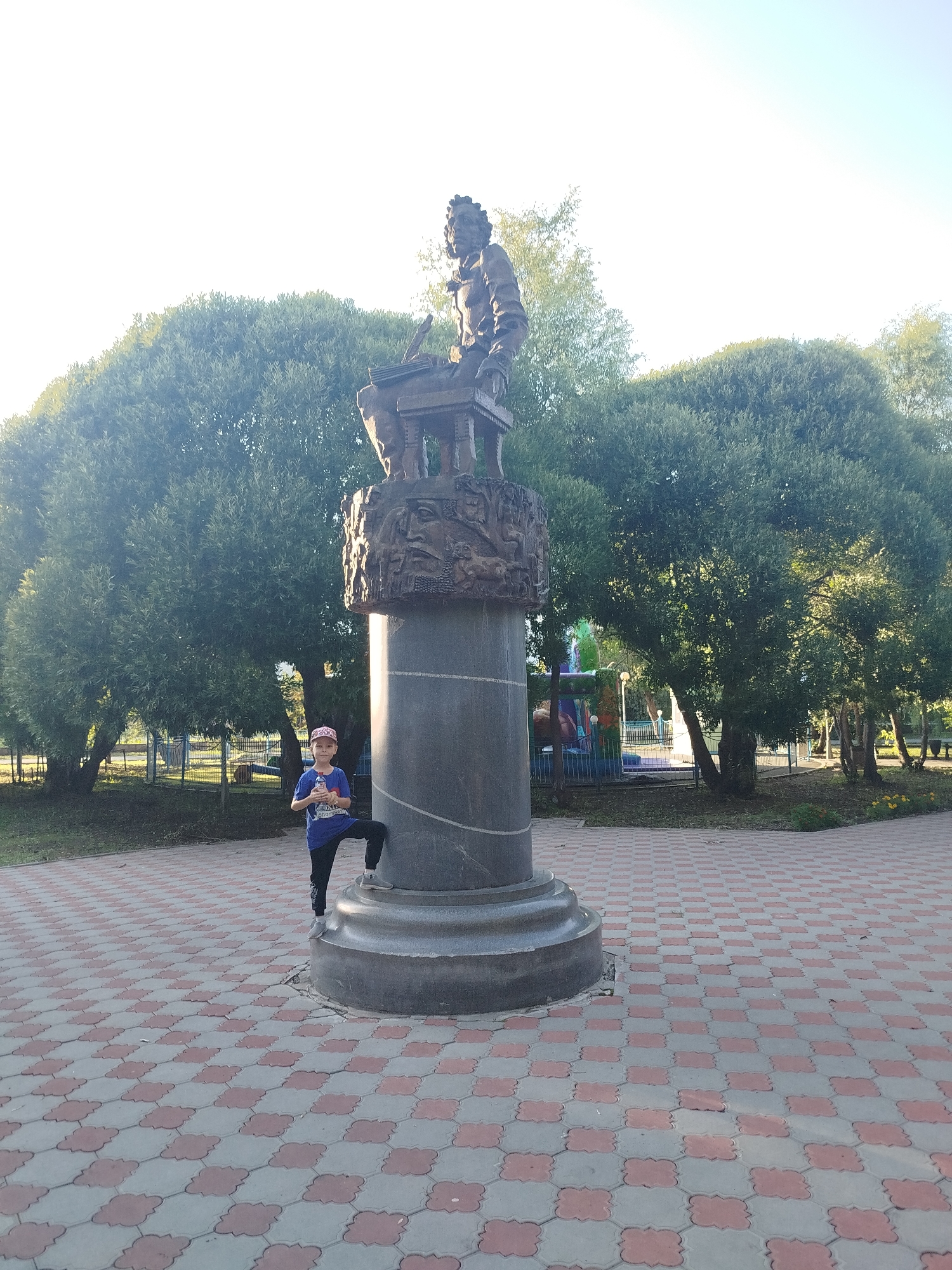 A young person wearing a blue shirt, black pants, and a pink cap stands next to a large monument in Yalutorovsk, Russia. The monument features a dark stone pedestal topped with a bronze sculpture of a seated figure at a desk, with intricate carvings around the base. The area is paved with a red and gray interlocking tile pattern. Lush green trees surround the monument, and a playground structure is visible in the background. The sky is clear and bright, suggesting a sunny day.