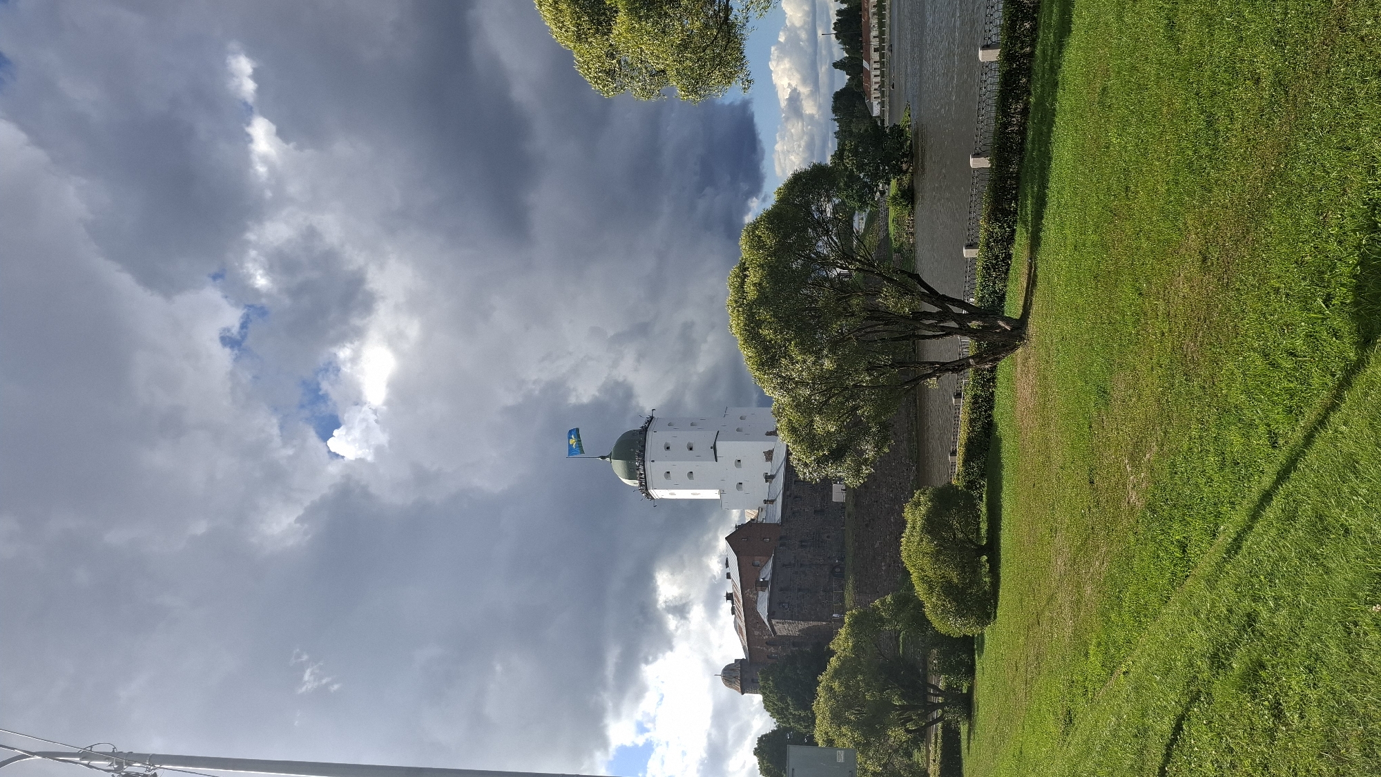 The image depicts Vyborg Castle, a medieval stronghold in Vyborg, Russia, under a dramatic, cloudy sky. The main structure is a tall, white tower with a green-domed roof, topped by a flag. Adjacent to it are older, stone fortifications and buildings. In the foreground, a well-maintained green lawn slopes down towards a body of water, likely the Vyborg Bay. Trees, some with full green foliage, are scattered across the landscape. A decorative fence runs along the edge of the lawn near the water. The lighting suggests it's daytime, possibly late afternoon, with pockets of sunlight breaking through the heavy clouds. There are no people visible in the frame.
