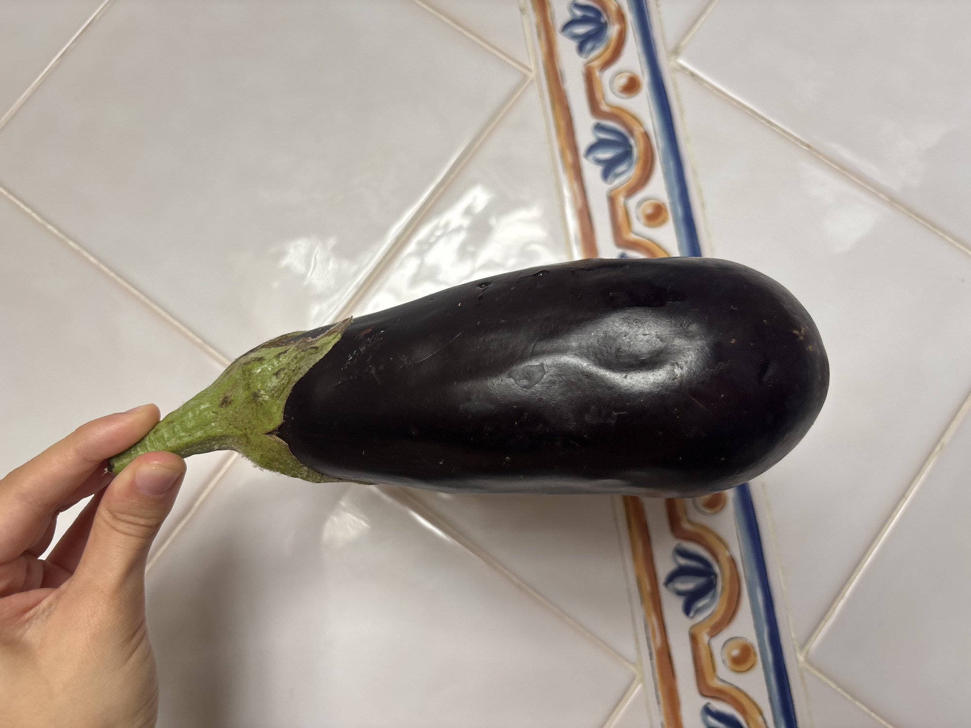 A hand is holding an eggplant in front of a tiled wall. The eggplant is dark purple and has a green stem. The wall is white and has a decorative border with a blue and orange pattern. The image appears to be taken indoors, possibly in a kitchen. The location in the city of Orihuela, Spain is unknown.