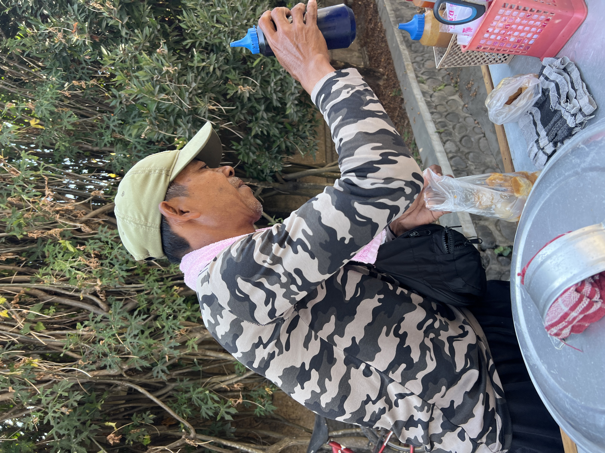 In Denpasar, Indonesia, an outdoor food vendor, a man wearing a camouflage long-sleeved shirt and a light green cap, is preparing food. He holds a dark blue squeeze bottle with a blue cap in his right hand, poised over a table. A pink towel is draped around his neck. On the table, there are several items: a clear plastic bag containing food items, a small plastic bag with more food, a folded checkered cloth, and a large silver circular object with a red and white striped cloth on it. In the background, there is dense green foliage and a low stone wall. To the right of the vendor, there are two condiment bottles and a pink plastic basket filled with packaging.