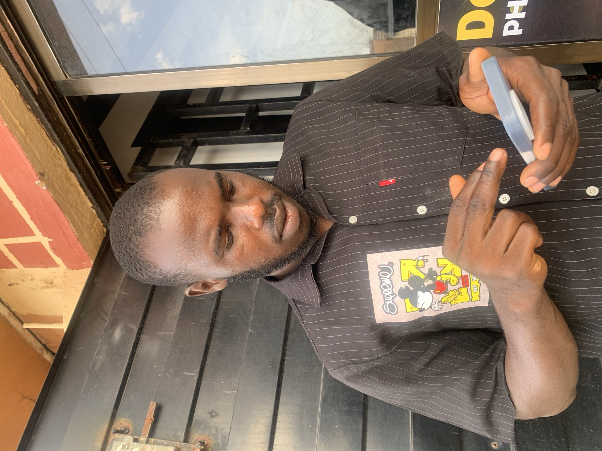 The image shows a man holding a blue and grey object near a window. He appears to be outdoors, near a building with a black facade. The man is wearing a brown, striped shirt with a cartoon patch on the chest. The environment is urban, with the possibility of the location being in Jos, Nigeria. No specific action or interaction can be definitively discerned from the image.