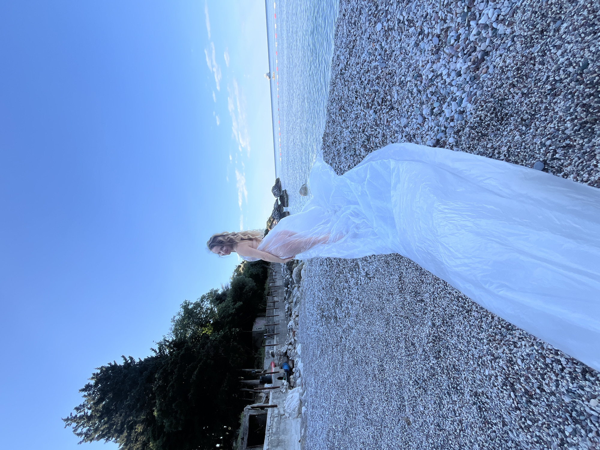 A partially clothed, light-skinned woman with long blonde hair stands on a pebbled beach in Boreti, Montenegro, partially covered in a large, white plastic sheet. The beach is on the edge of the Adriatic Sea under a clear blue sky with scattered clouds. A line of buoys stretches across the water. Behind the woman, there's a concrete retaining wall with a damaged or abandoned building, partially obscured by trees.