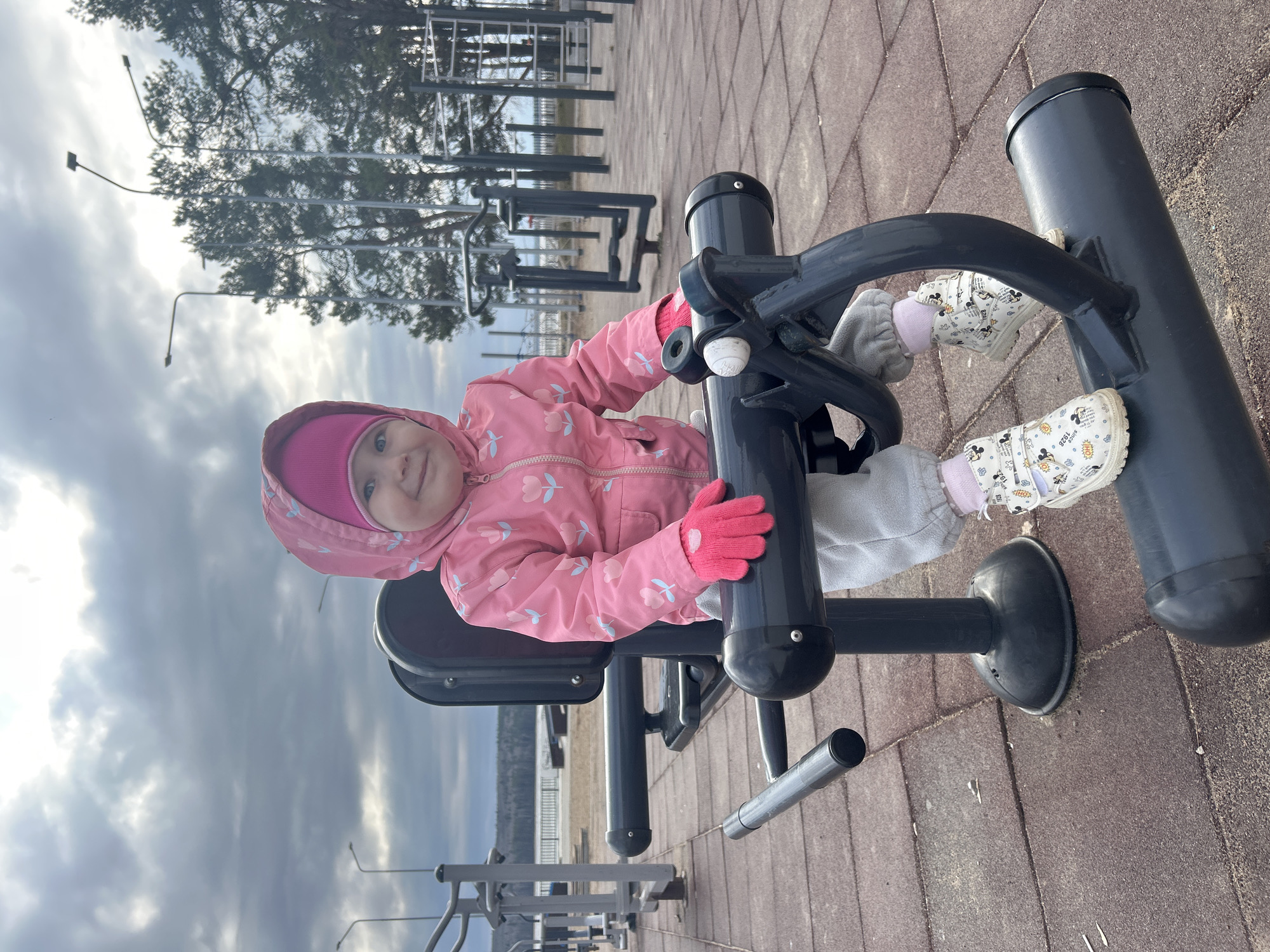 A young child wearing a pink jacket, matching hat and gloves, and white shoes is sitting on an outdoor exercise machine. The machine is black and has handlebars that the child is holding. The child is smiling and looking at the camera. The location is an outdoor fitness area in Chaikovsky, Russia, identifiable by the lake visible in the background and the typical layout of the exercise area. The ground is covered in red brick paving. Other exercise equipment is visible in the background. The sky is overcast.