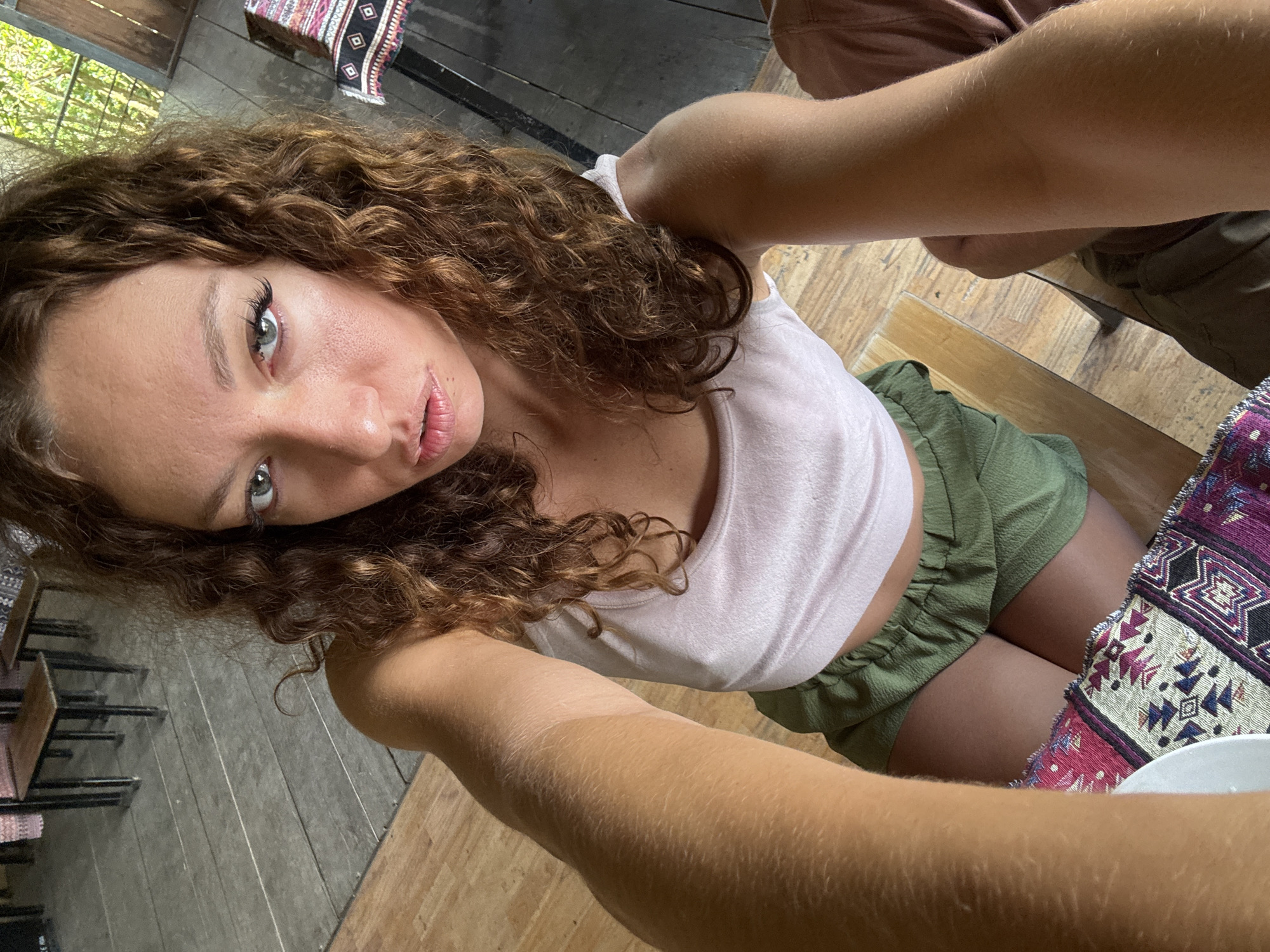 A selfie shot from a slightly elevated angle showcases a woman with curly brown hair, wearing a light pink tank top and green shorts. She is situated indoors, with a gray-toned floor to her left and light brown wooden flooring beneath her. To the right, a colorful patterned object sits on a table. Based on the surroundings, including furniture style, and the context of Ngã Ba Kim, Vietnam, it can be inferred that the location is likely a cafe or restaurant. The woman is holding the camera, her arms visible in the foreground.