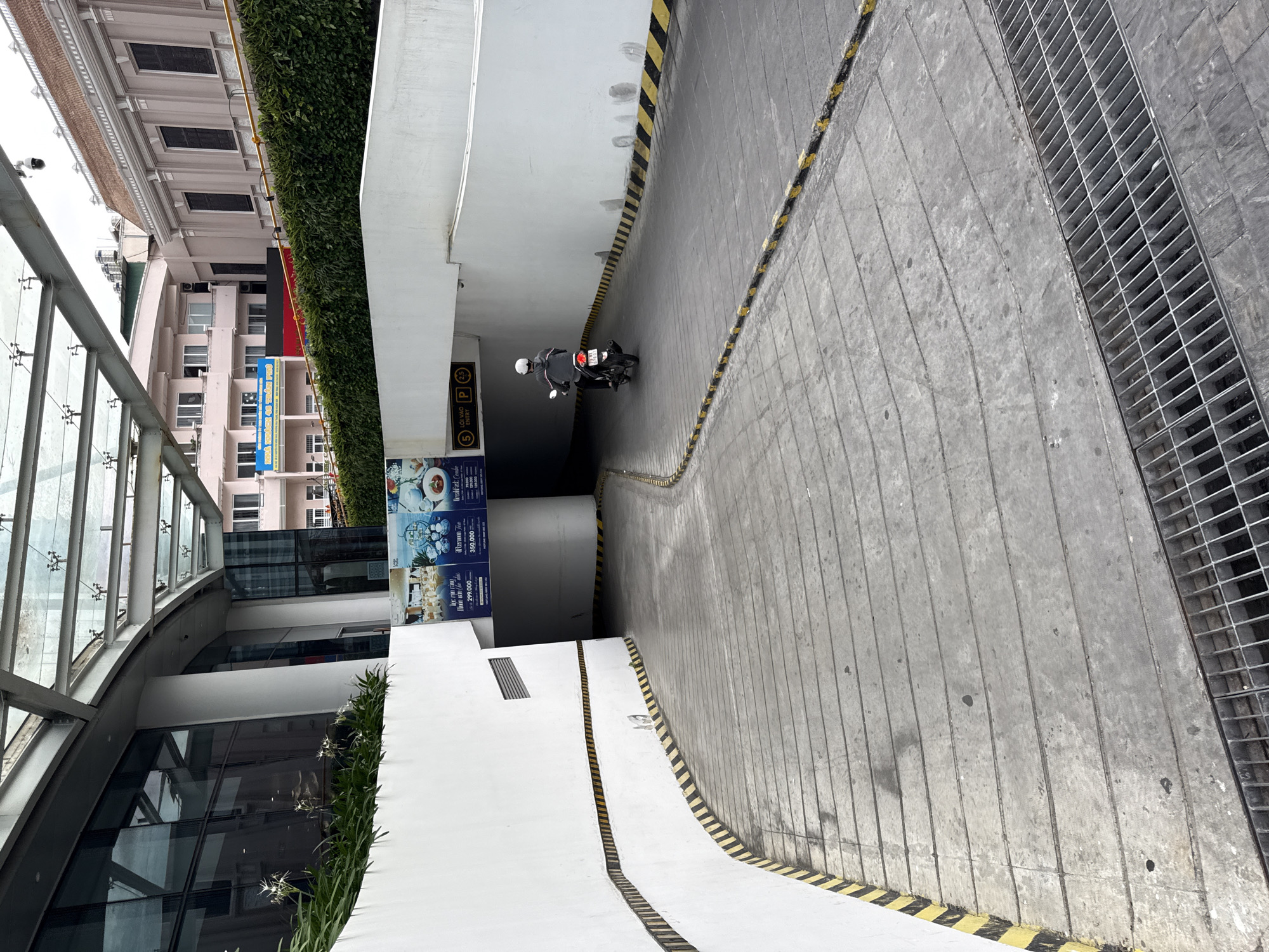 This image depicts the entrance to an underground parking garage in Nha Trang, Vietnam. A concrete ramp leads down into the garage, bordered by white walls with alternating yellow and black hazard stripes. A person is riding a motorcycle down the ramp. Above the entrance, a large banner advertises dining specials. The surrounding buildings suggest a commercial or mixed-use urban environment.