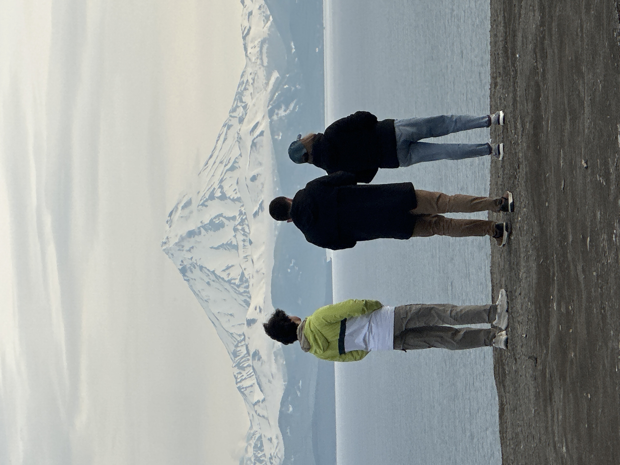 Three individuals stand on a rocky, dark-colored shore, their backs to the camera, gazing out at a body of water. Beyond the water, a large, snow-capped mountain dominates the horizon, its peak shrouded in soft, cloudy skies. The person on the left wears a bright green and white hooded jacket and tan cargo pants. The person in the center is dressed in a dark jacket and brown pants. The person on the right wears a dark jacket, light blue jeans, and a baseball cap. The scene is likely overlooking the Avacha Bay, with the iconic Koryaksky or Avachinsky volcanoes in the background, a common vista from Petropavlovsk-Kamchatsky, Russia.