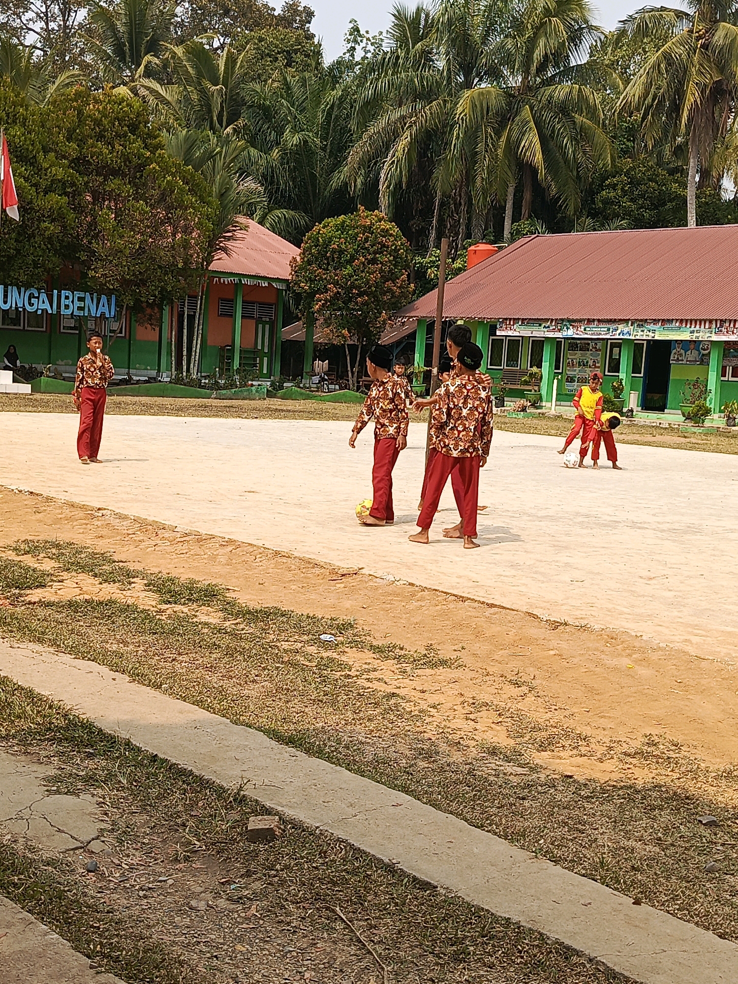 In an outdoor courtyard, children play soccer in front of a school building with a red-tiled roof and green walls. The school has a sign that reads "GUNGAIBENAI" in blue letters. Palm trees and other foliage surround the school. Several children, dressed in patterned shirts and red pants, are visible in the image. Two boys are actively engaged in playing soccer, one with his foot on the ball and the other bending down. Another boy stands to the left, watching the game, while a fourth boy stands further to the right. The ground is a light brown dirt or sand, with patches of dry grass. The sky is bright and hazy, suggesting a sunny day.
