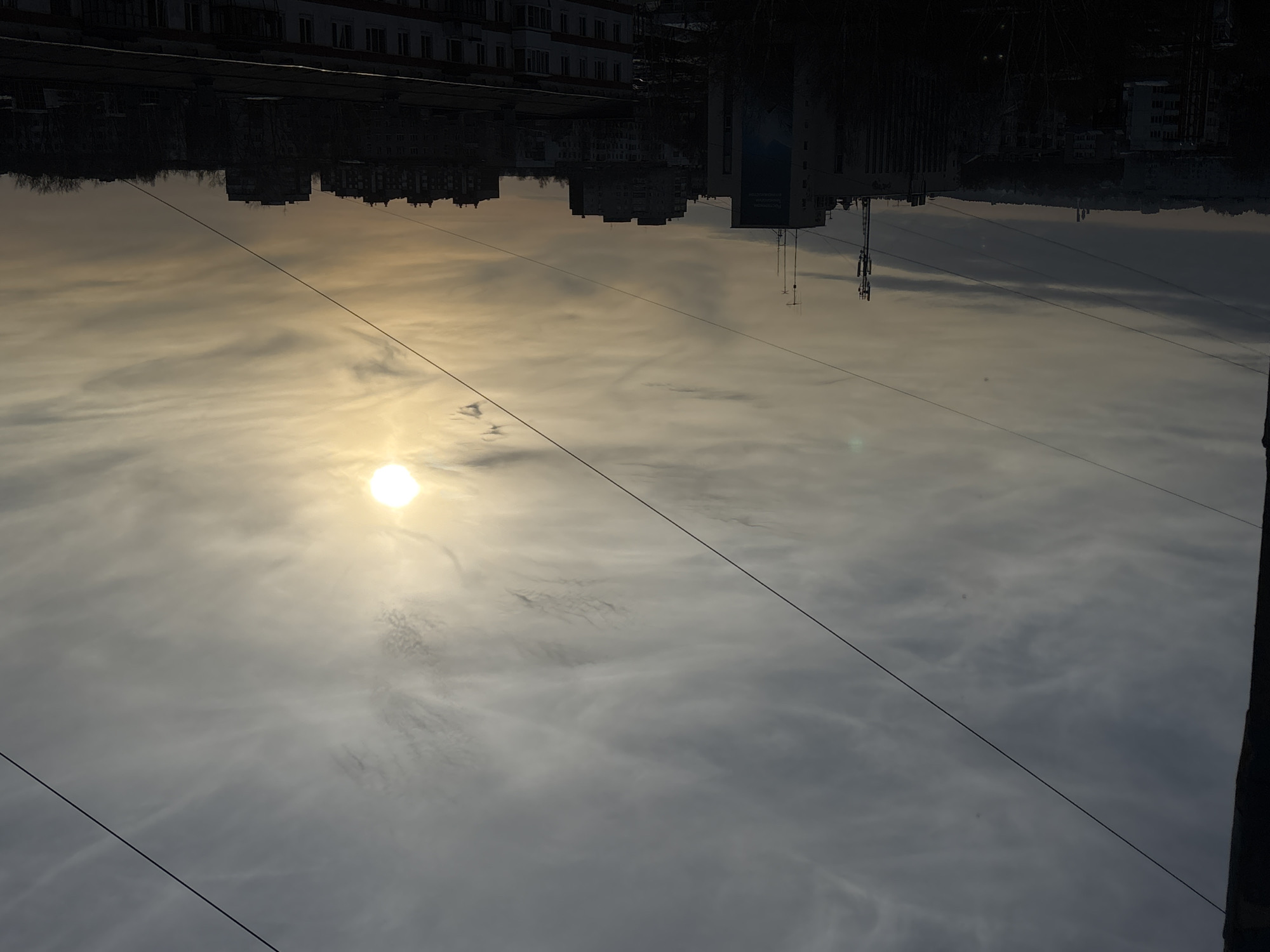 The image presents an elevated urban landscape view of Chaikovsky, Russia, under a partly cloudy sky with a low sun, indicating either late afternoon or early morning. The scene is predominantly backlit, causing most foreground and midground elements to appear in silhouette.
The upper two-thirds of the frame are dominated by the sky, filled with streaky and patchy cirrus and stratocumulus clouds. The sun, a bright white-yellow orb, is prominent in the upper right quadrant, partially obscured by thin cloud cover, casting a warm, diffuse illumination. The sky's color palette ranges from light grey-blue in the upper left to warm yellow and orange around the sun, fading to darker grey tones near the horizon. Two distinct dark, thin utility lines or cables diagonally traverse the sky from the upper left to the lower right, with additional faint lines visible horizontally in the upper right.
The lower third of the image features a dense cityscape composed of multi-story buildings. In the midground, a light-colored building with a large blue advertisement or mural on its side is visible, its rooftop equipped with multiple communication antennas and cellular masts. Surrounding this are numerous residential apartment blocks, appearing as dark, indistinct silhouettes against the brighter sky, characteristic of Soviet-era architecture. Sparsely leaved or bare trees are interspersed among the buildings, also in silhouette. On the distant horizon, a low, vegetated ridge is visible. Faintly discernible against the sky on the central-left horizon are two tall, industrial-like structures, possibly chimneys or cooling towers, consistent with the infrastructure of the Votkinsk Hydroelectric Power Plant in the Chaikovsky region. No individuals, specific actions, or interactions are present in the scene.