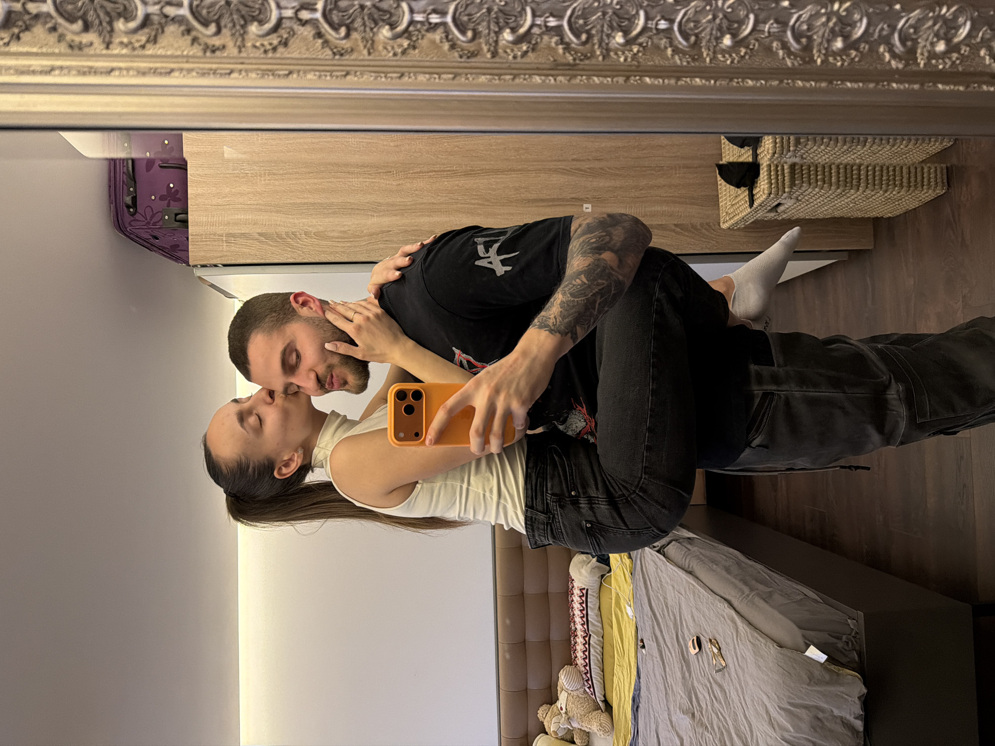 A reflection in an ornate, rectangular mirror shows a couple in a bedroom. A man in a black t-shirt and dark jeans embraces a woman in a white tank top and black jeans. They appear to be kissing as the woman holds up a mobile phone with an orange case, seemingly taking a selfie. In the background, there is a light-colored cabinet, a wicker basket on top of it, and a bed with gray bedding. Part of a headboard can be seen behind the bed, along with a stuffed teddy bear. There is also a suitcase with a floral pattern on top of the cabinet. The location appears to be an interior setting, such as a bedroom, but there is no indication of Sofia, Bulgaria.