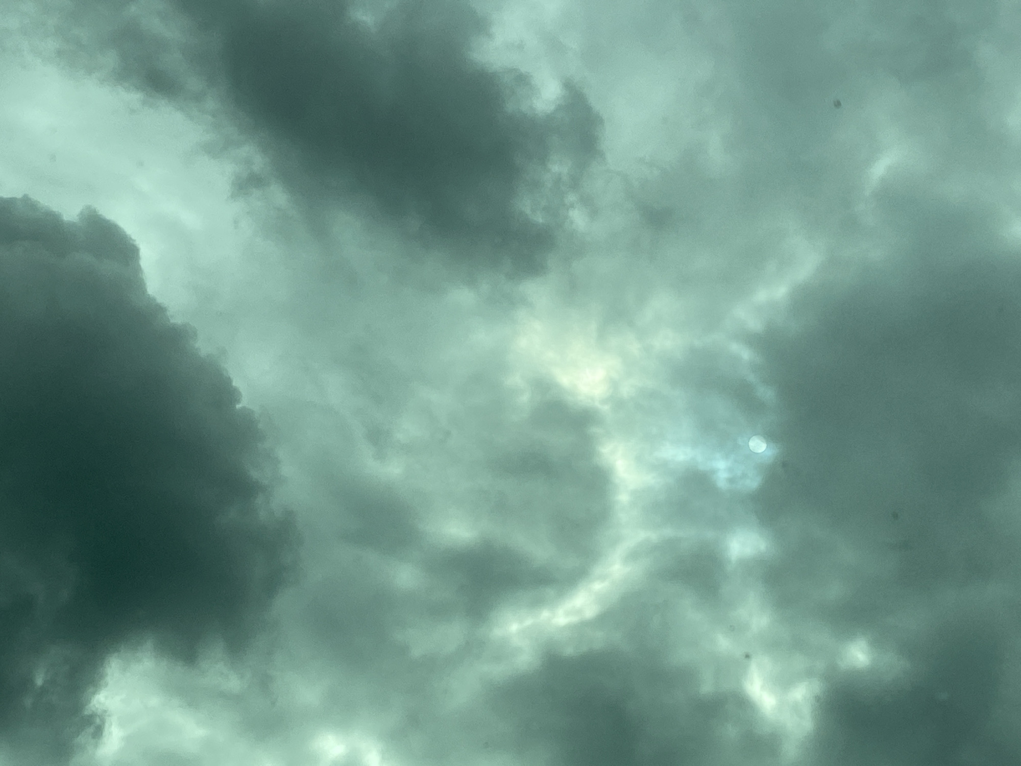 The image displays an overcast sky dominated by a thick expanse of clouds. The clouds vary in density, with darker, heavier formations appearing in the bottom left and top right sections, suggesting a deep, stormy, or pre-storm atmosphere. Lighter, more translucent clouds are visible in the central and middle-left areas.
A prominent feature is a bright, circular light source, likely the sun, partially obscured by a thin patch of clouds in the upper-right-middle of the frame. This light creates a luminous glow behind the lighter cloud layers, contrasting with the darker masses.
The overall color palette is a muted greenish-grey or teal, contributing to a moody and somewhat ominous impression. There are no identifiable people, objects, or visible text. The setting is solely the sky, providing no specific location cues. Small, dark, fuzzy specks are scattered across the image, suggesting dust on the camera lens or sensor. The weather appears heavily cloudy, and the light indicates it is likely daytime, despite the obscured sun.