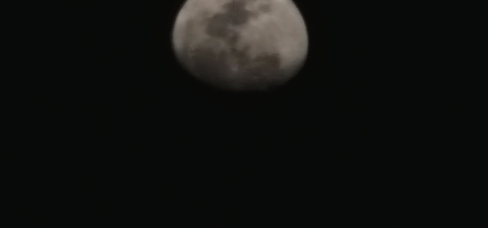 A partially illuminated moon is visible in a dark, starless sky. The moon is positioned in the right half of the frame, appearing as a sphere with visible craters and maria, suggesting a gibbous phase. The left side of the image is predominantly black, creating a stark contrast with the celestial body. No other objects or environmental features are discernible. The location is unknown.